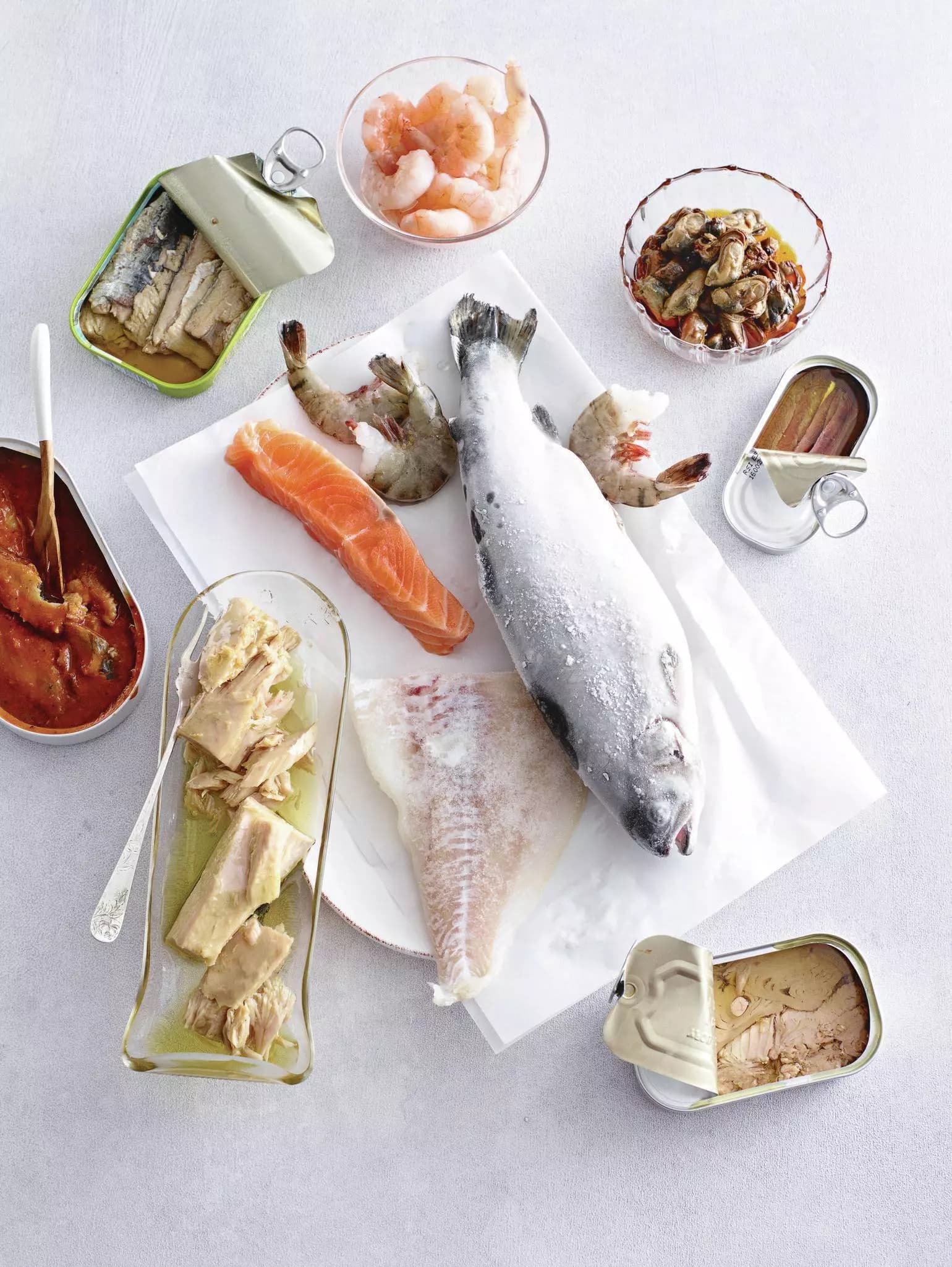 Auf dem Bild sieht man verschiedene Fischprodukte und Meeresfrüchte, darunter eine Dose Sardinen, Garnelen, Lachs, eine Dose Sardellen, Muscheln, eingelegter Fisch, ein Fischfilet und eine Dose Thunfisch.