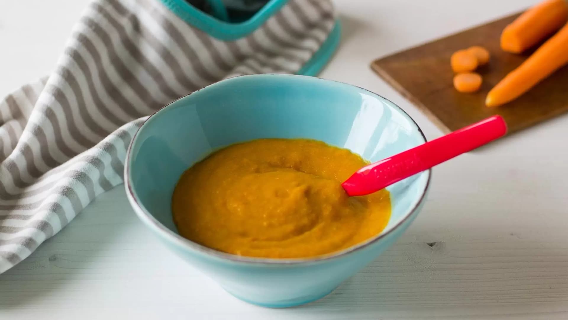 Schale mit orangenem Brei und rotem Löffel auf weißem Tisch neben gestreiftem Geschirrtuch und Holzbrett mit Karottenstücken.