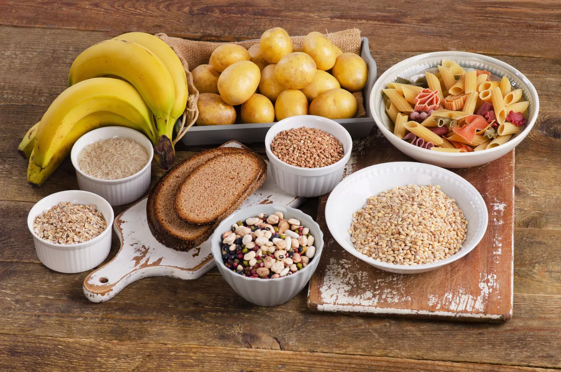 Ein Ensemble kohlenhydrathaltiger Lebensmittel wie Bananen, Haferflocken, Brot, Kartoffeln, Nudeln und Reis angerichtet auf Holz.