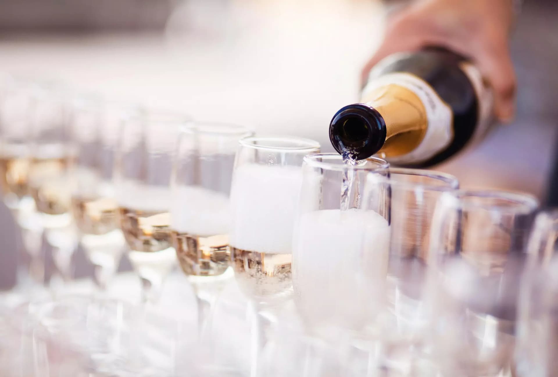 Eine Hand schenkt Schaumwein aus einer klassischen grün-goldenen Flasche in viele, aneinandergereihte Sektgläser ein