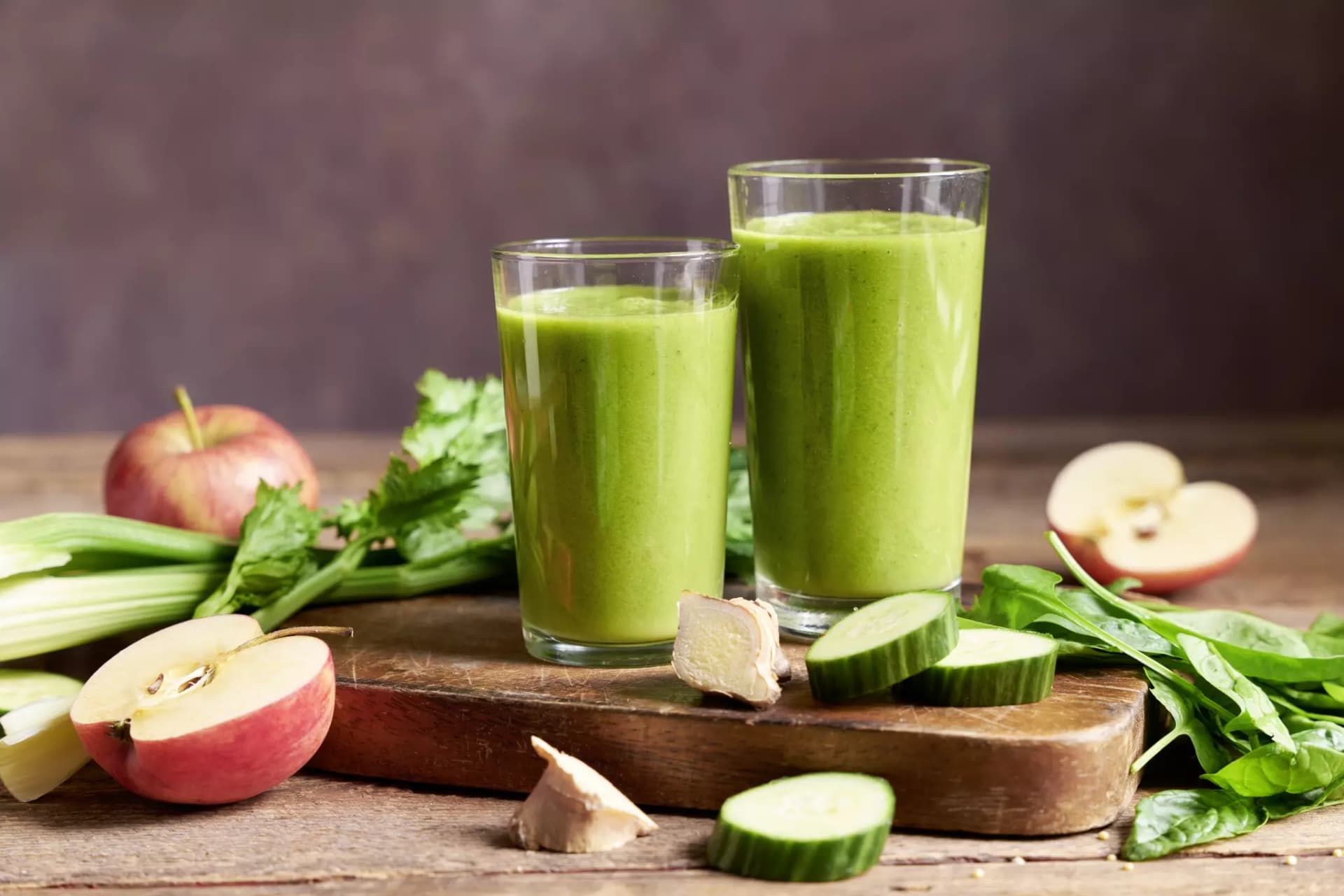  Zwei Gläser mit grünem Smoothie auf einem Holzbrett umgeben von Apfelhälften, Gurkenscheiben, Ingwerstücken und grünen Blättern.