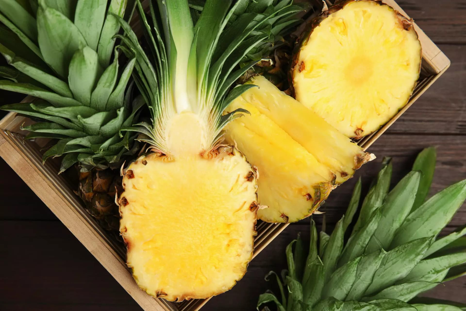 Eine zerteilte Ananas liegt auf weiteren, unzerteilten Ananas in einem rechteckigen Holzkorb.
