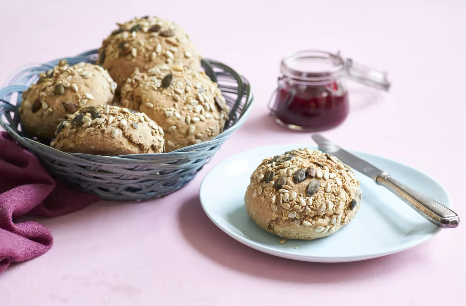 Brötchen mit verschiedenen Kernen in einem Korb und auf einem Teller mit Messer und kleinem Glas mit roter Marmelade auf rosa Untergrund.
