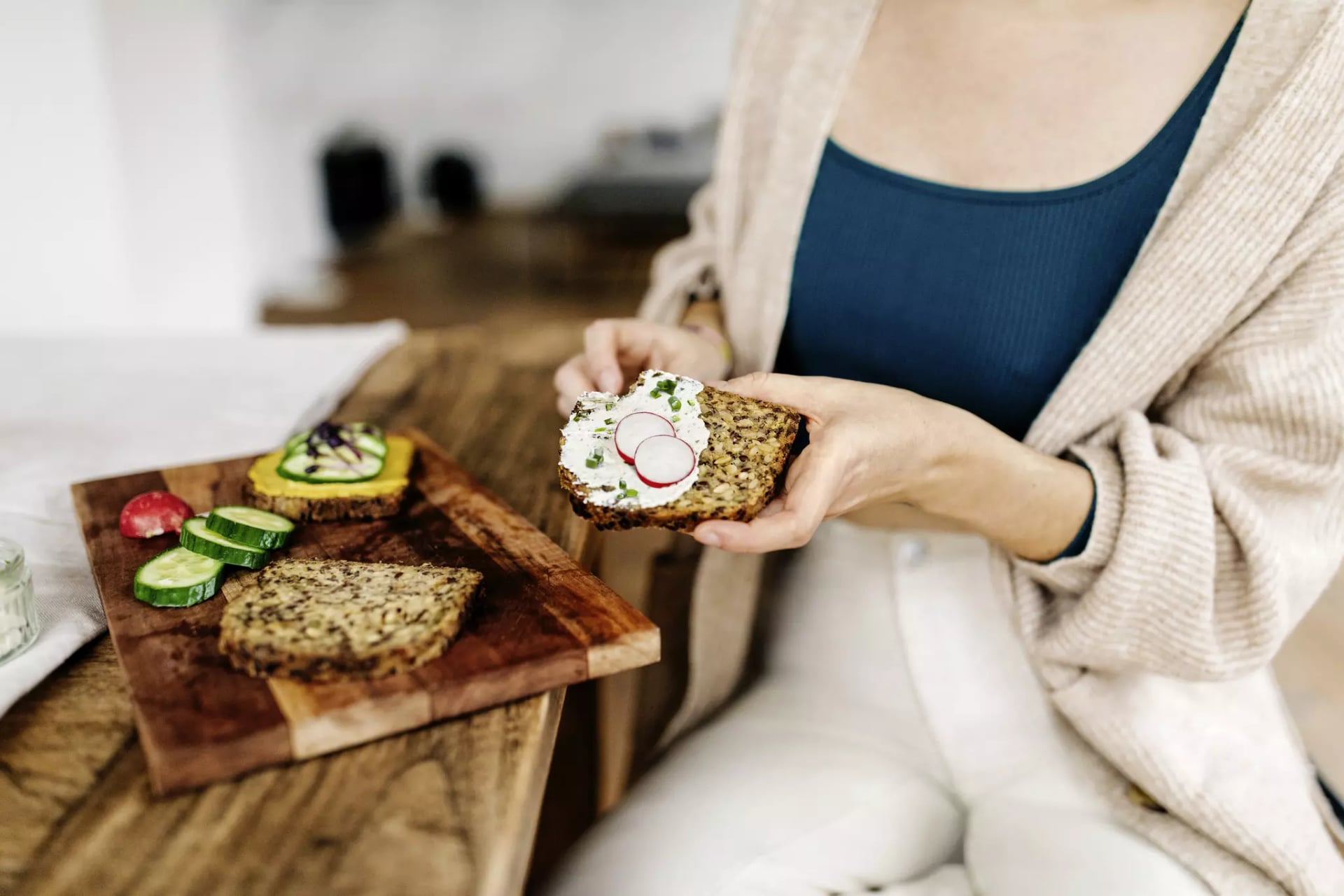 Eine Person sitzt an einem Holztisch. Vor ihr steht ein Brett auf dem verschiedenes Gemüse, Aufstriche und Scheiben von glutenfreiem Brot liegen.