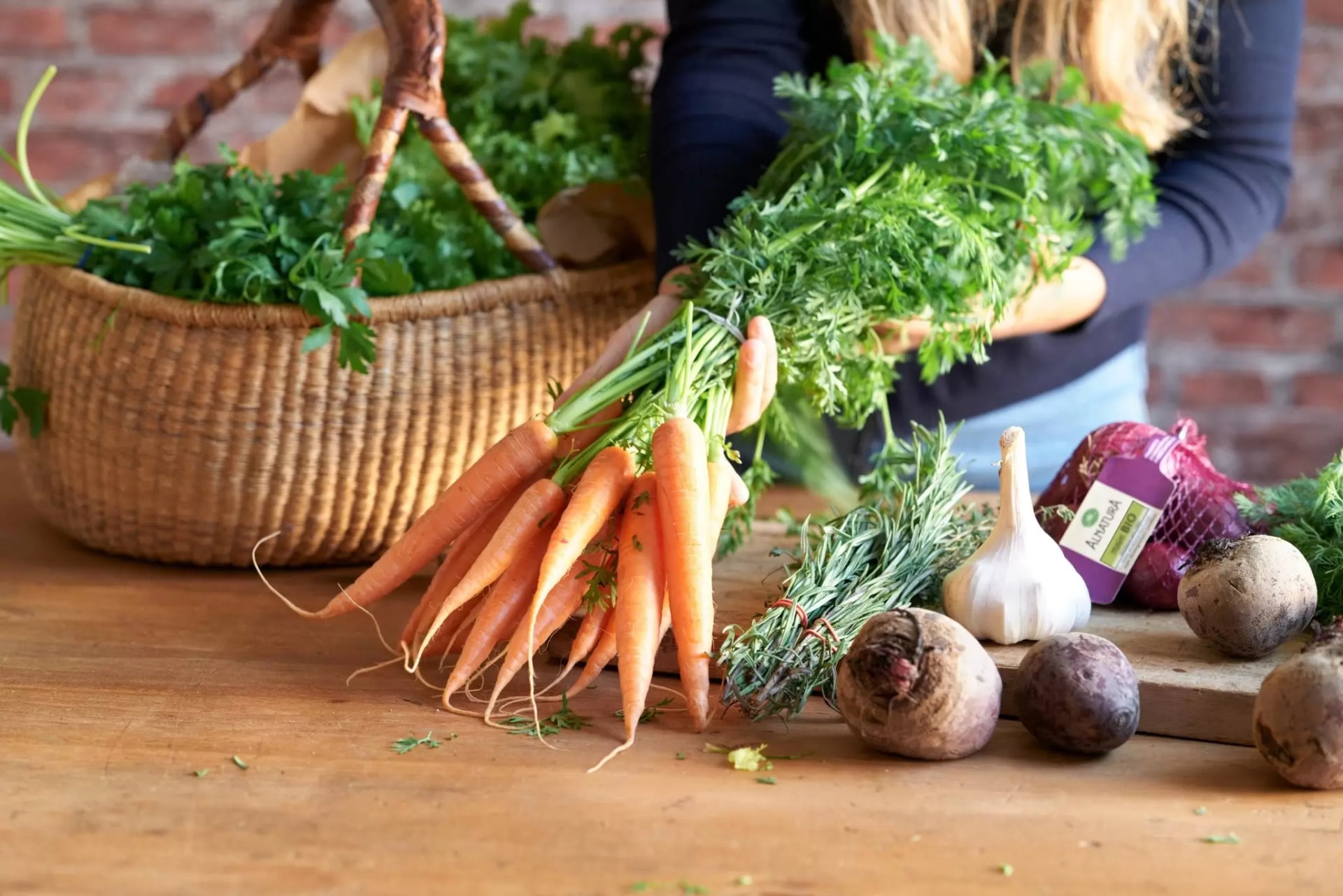 Person hält ein Bündel frischer Karotten über einem Holztisch mit Knoblauch, Rote Bete und einem Korb mit grünem Blattgemüse im Hintergrund.
