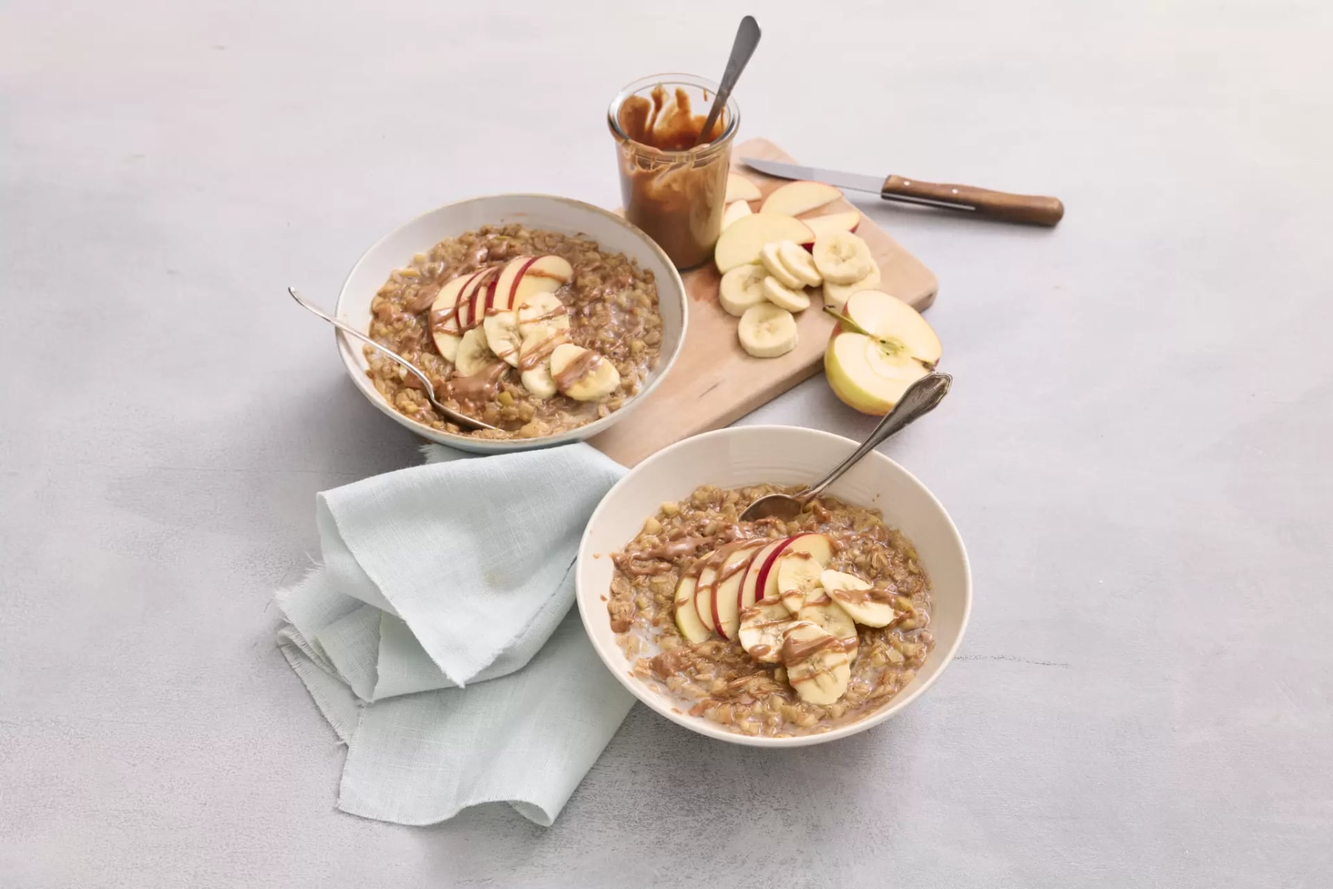 In zwei weißen Porzellanschüsseln sind Porridge garniert mit Apfelscheiben und Mandelmus. Seitlich in der Schüssel steckt ein Teelöffel. Im Hintergrund liegt eine hellblaue Stoffserviette und ein Holz-Schneidebrett auf dem das Mandelmus und Bananen- und Apfel-Scheiben liegen.