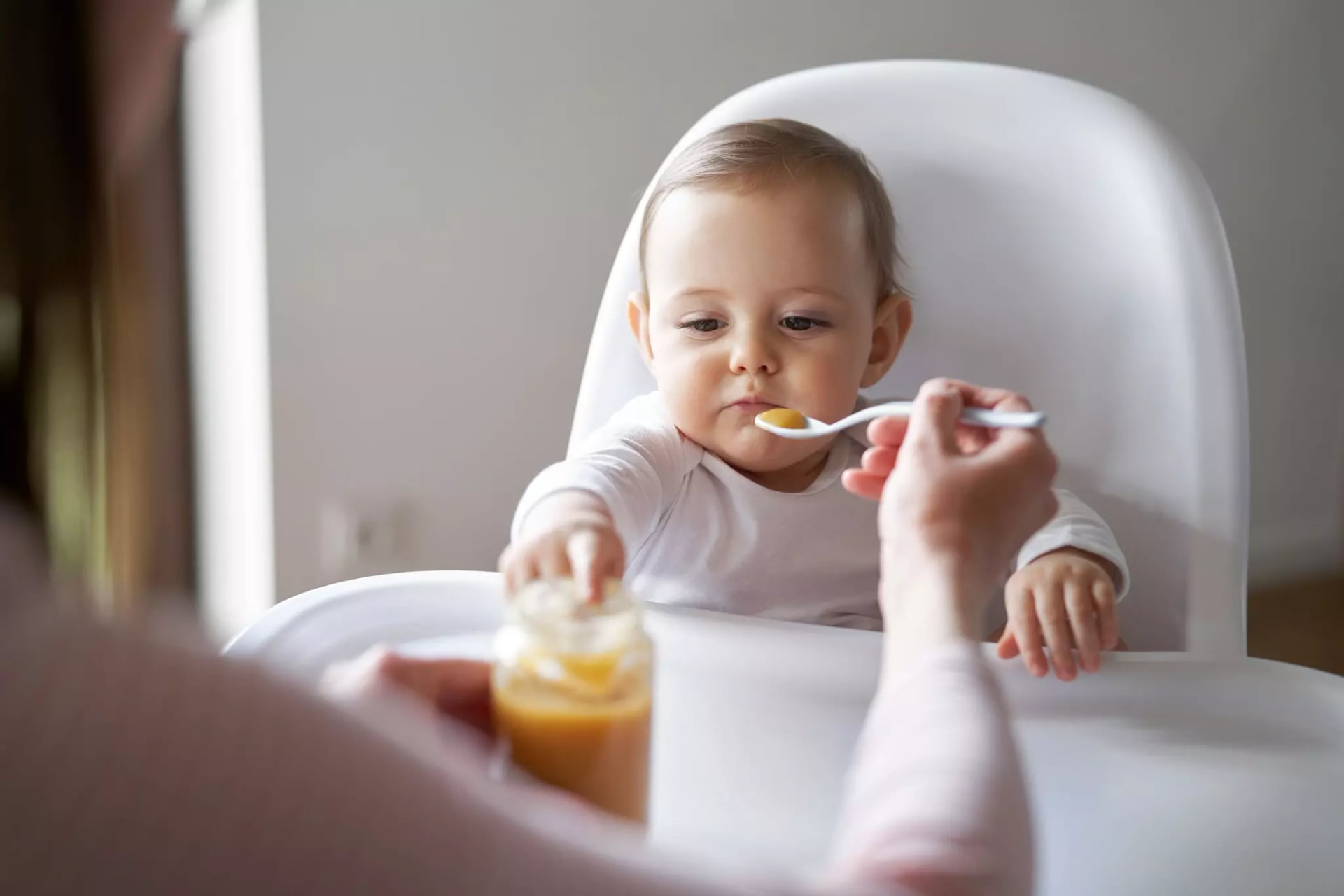 Ein Baby sitzt auf dem Hochstuhl und wird mit einem weißen Löffel von einer Frau gefüttert. Es greift nach dem Brei im Glas.