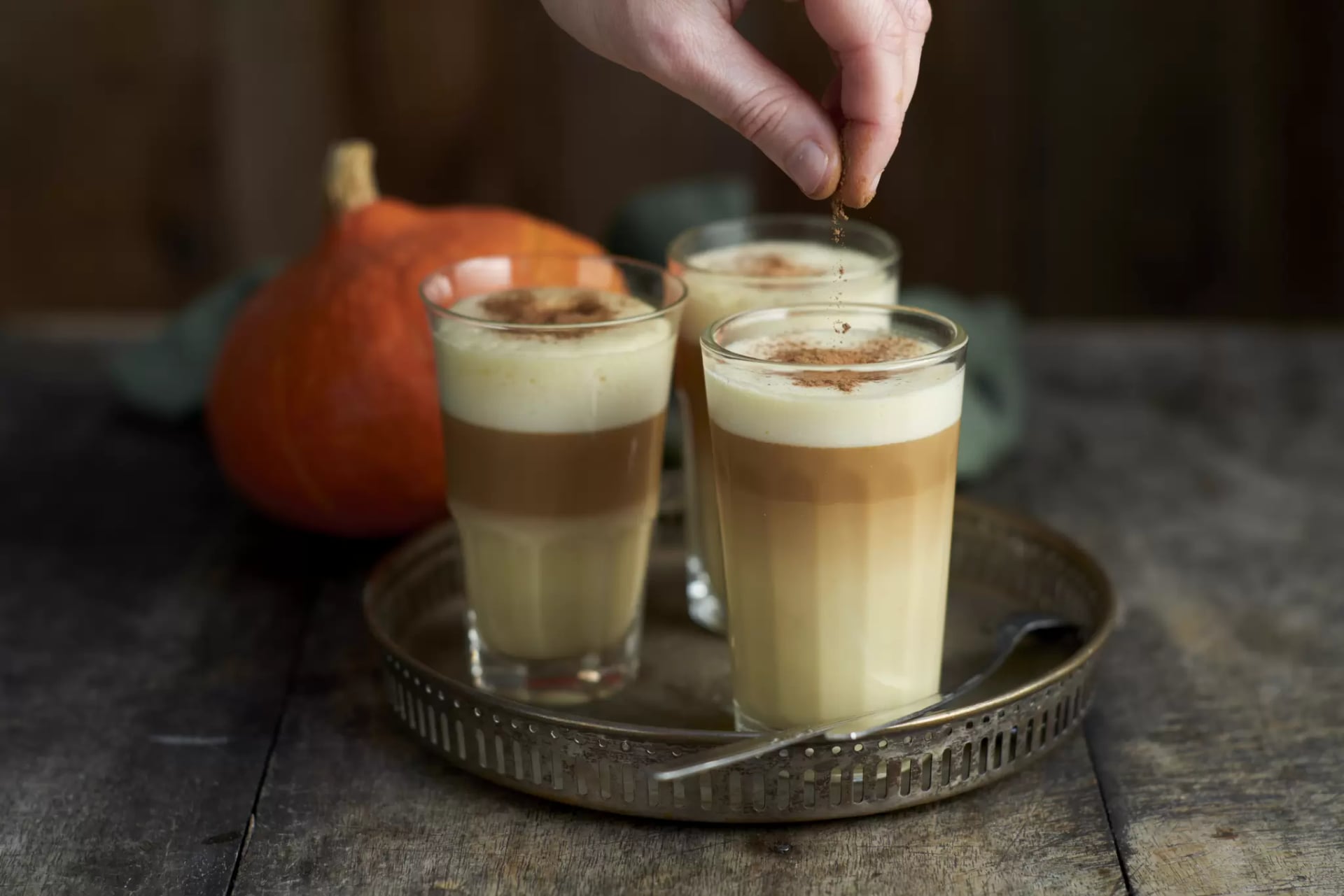Drei hohe Gläser mit geschichteten Pumpkin Spice Latte, stehen auf einem runden, antiken Metalltablett. Die Getränke zeigen einen schönen Farbverlauf von hellem Gelb über Braun bis hin zu einer festen, weißen Schaumkrone. Eine Hand streut gerade eine Prise Zimt oder Kakaopulver auf den Milchschaum eines Glases. Im dunklen Hintergrund ist ein ganzer Hokkaido-Kürbis auf einem Holztisch zu sehen.