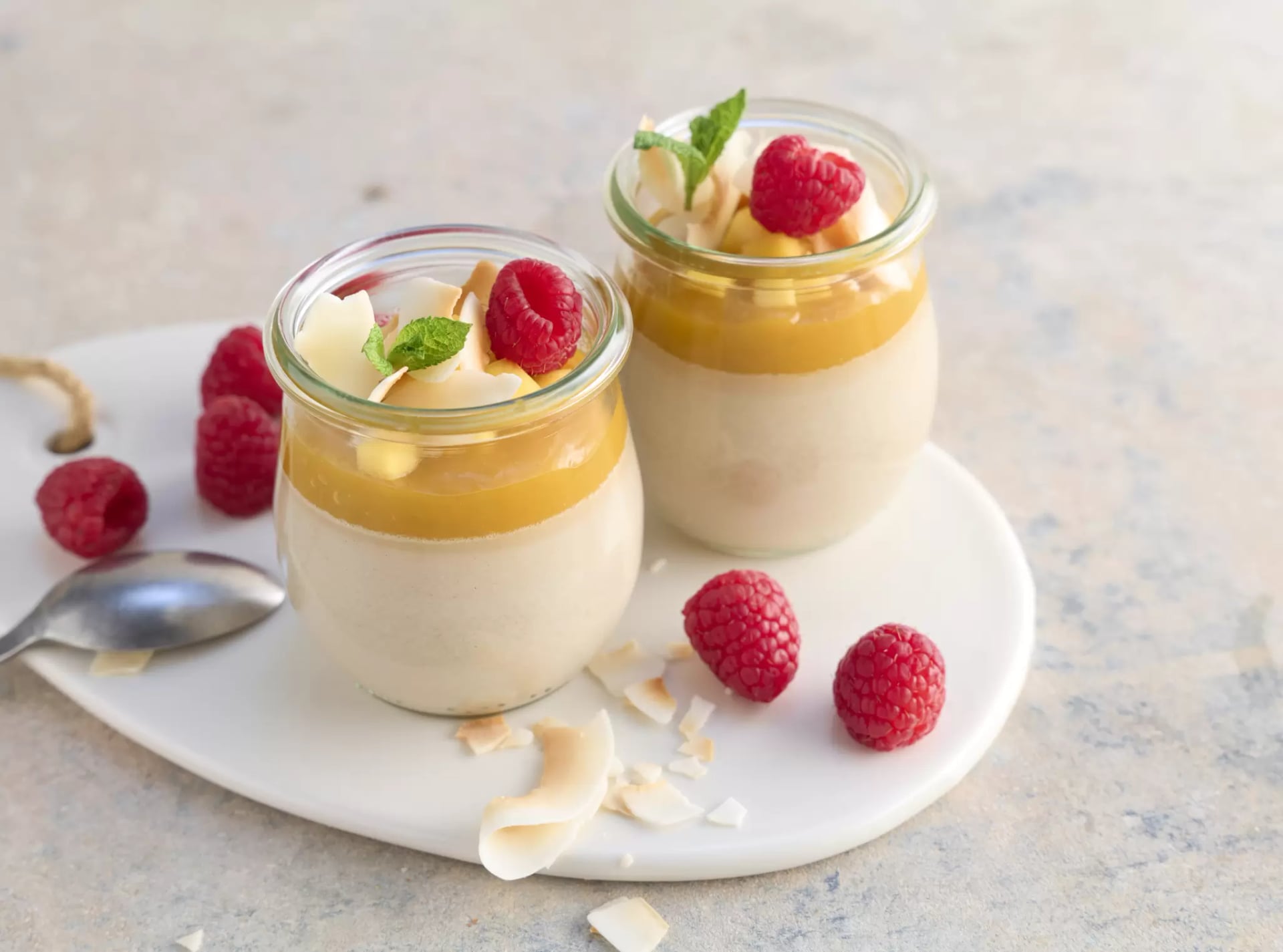 Zwei Gläser vegane Koko-Panna-cotta mit Mangosauce, Himbeeren, Kokoschips und Minzblättern auf weißem Teller mit einem Löffel am Tellerrand und weiteren Himbeeren.