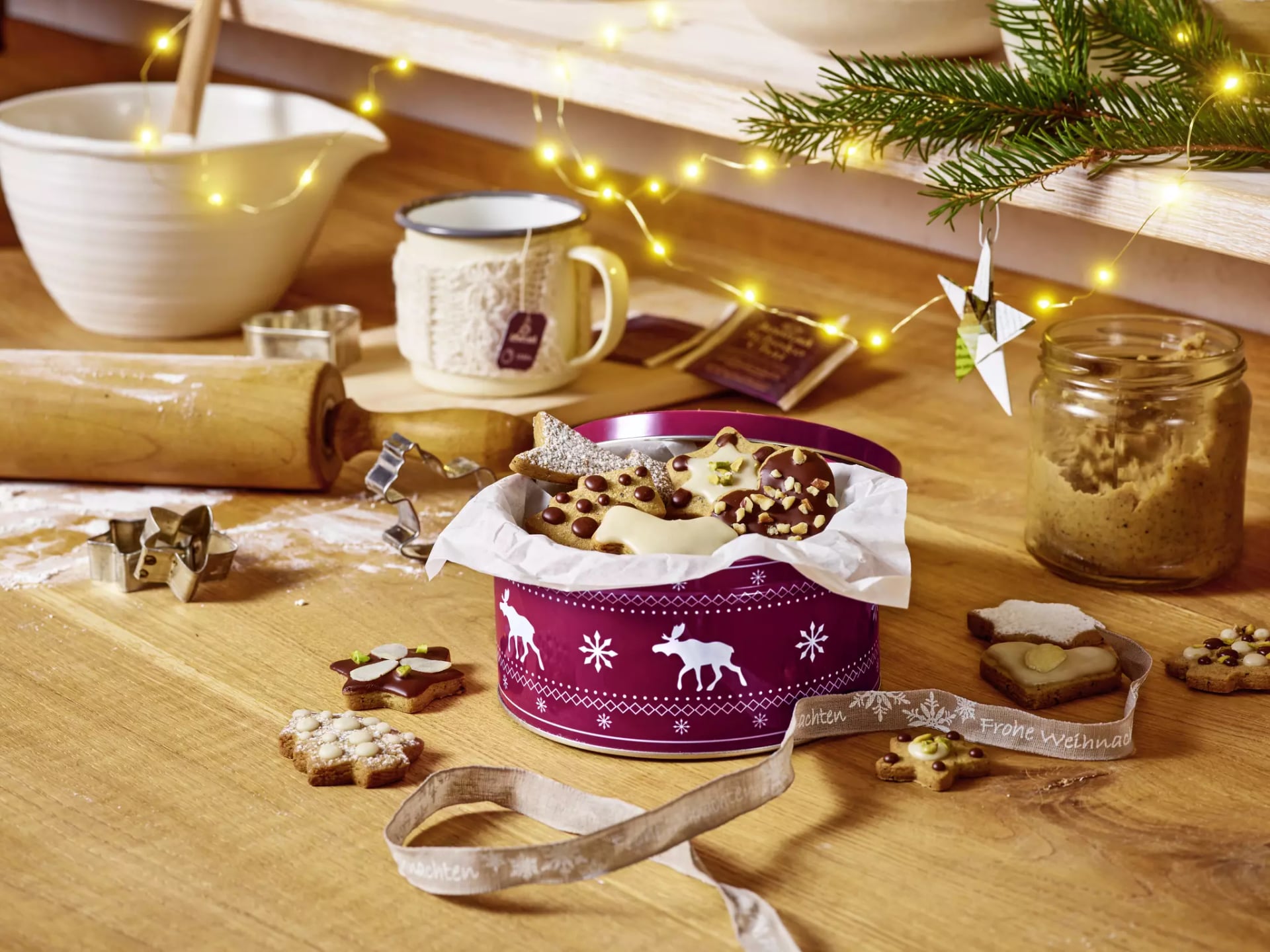Mit Plätzchen gefüllte Keksdose auf einem Holztisch mit Plätzchen-Ausstechern, Plätzchenteig und einer Tasse Tee.