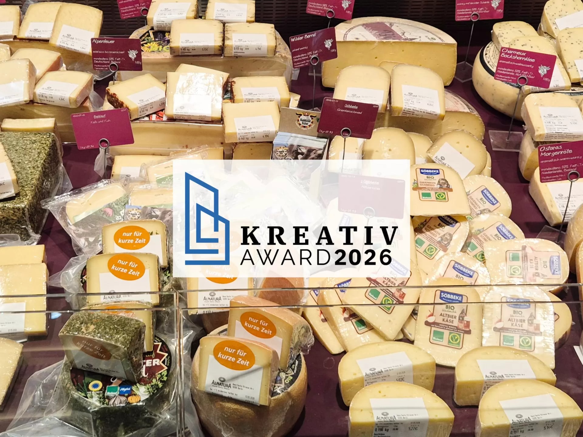 Logo des "Kreativ Award 2026" auf dem Bild einer gutbestückten Käsetheke mit verschiedenen abgepackten Käsespezialitäten.