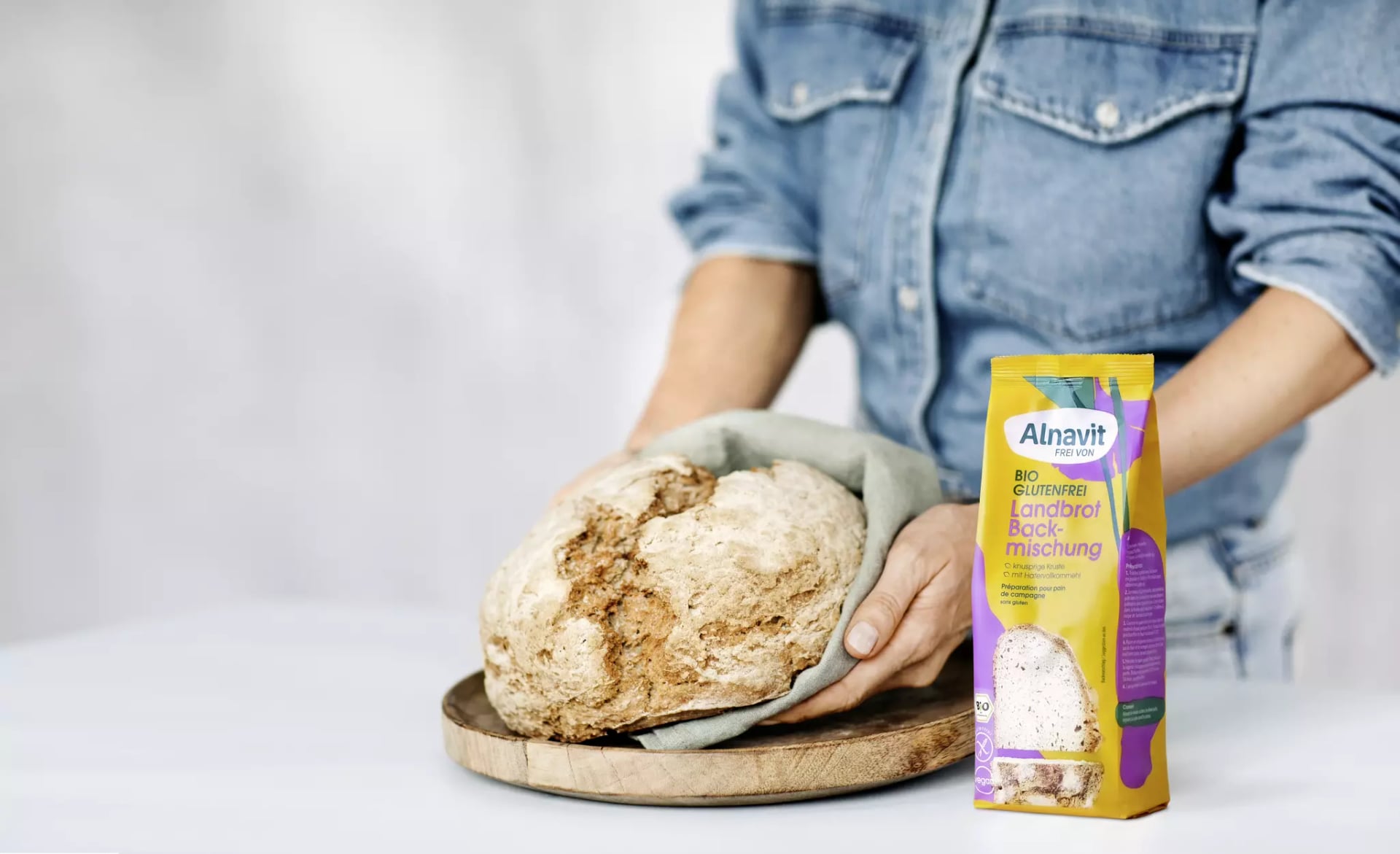 Eine Person legt ein glutenfreies Brot auf ein Brett. Dafür verwendet Sie ein Küchenhandtuch. Neben dem Brot steht die Landkorn-Backmischung der Marke Alnavit.