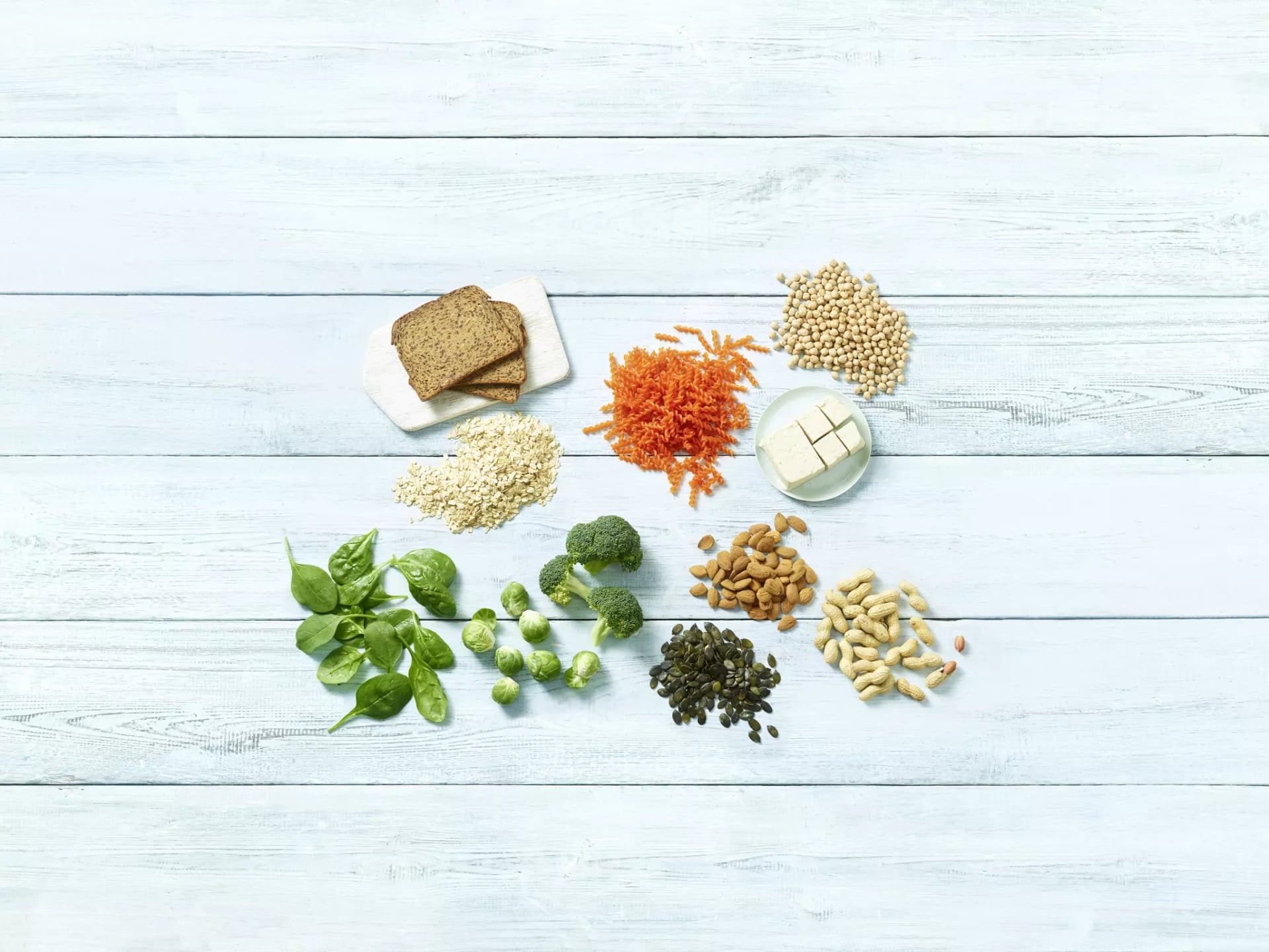 Verschiedene pflanzliche Lebensmittel wie Vollkornbrot, Haferflocken, geriebene Karotten, Kichererbsen, Tofu, Spinat, Rosenkohl, Brokkoli, Mandeln, Kürbiskerne und Erdnüsse auf hellem Holzhintergrund.