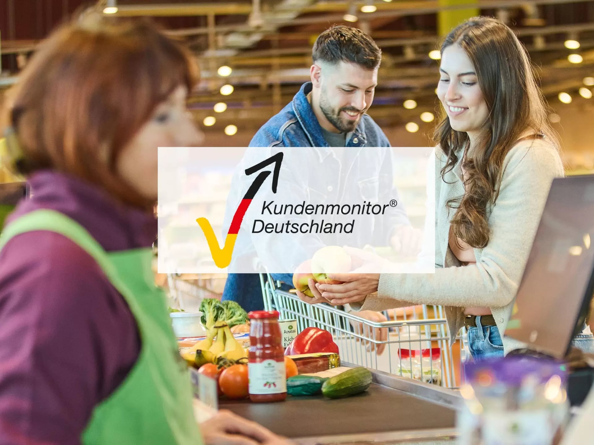 Einkaufssituation mit einer Frau und einem Mann an der Kasse eines Alnatura Marktes. Darüber ist das Logo von Kundenmonitor Deutschland zu sehen.