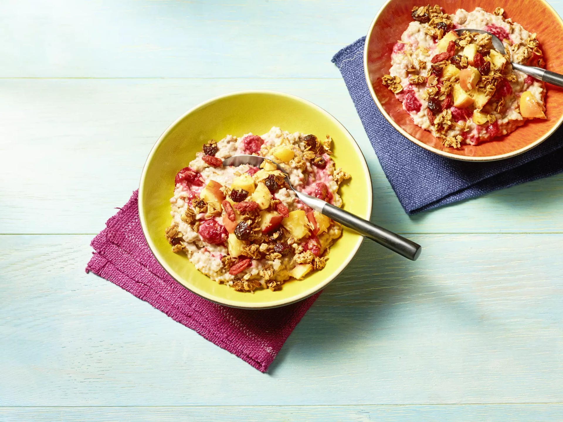Zwei Schalen mit Haferbrei, Obststücken und Granola, jeweils mit einem Löffel, auf farbigen Stoffunterlagen auf hellem Holztisch.
