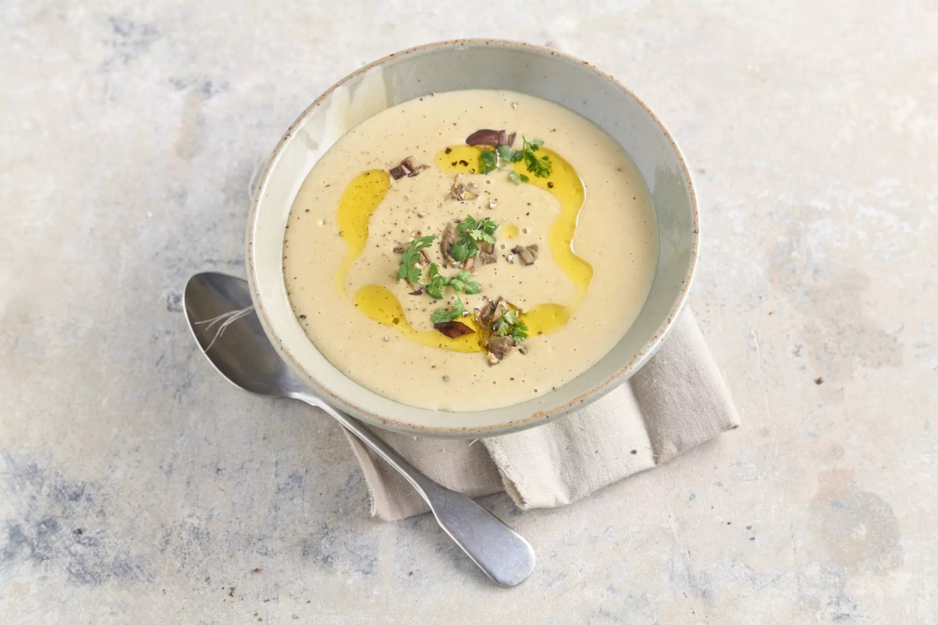 Schale mit cremiger Suppe, garniert mit Kräutern, Olivenöl und kleinen Pilzstücken, auf einem dunklen Tuch und einem rustikalen Holztisch