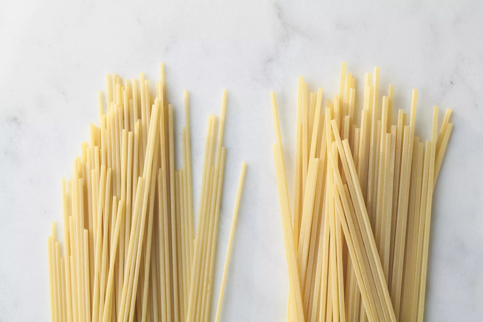 Ein Bündel ungekochter Spaghetti auf der linken Seite und ein Bündel ungekochte Spaghetii Quadrati auf der linken Seite, beides auf weißer Marmoroberfläche.