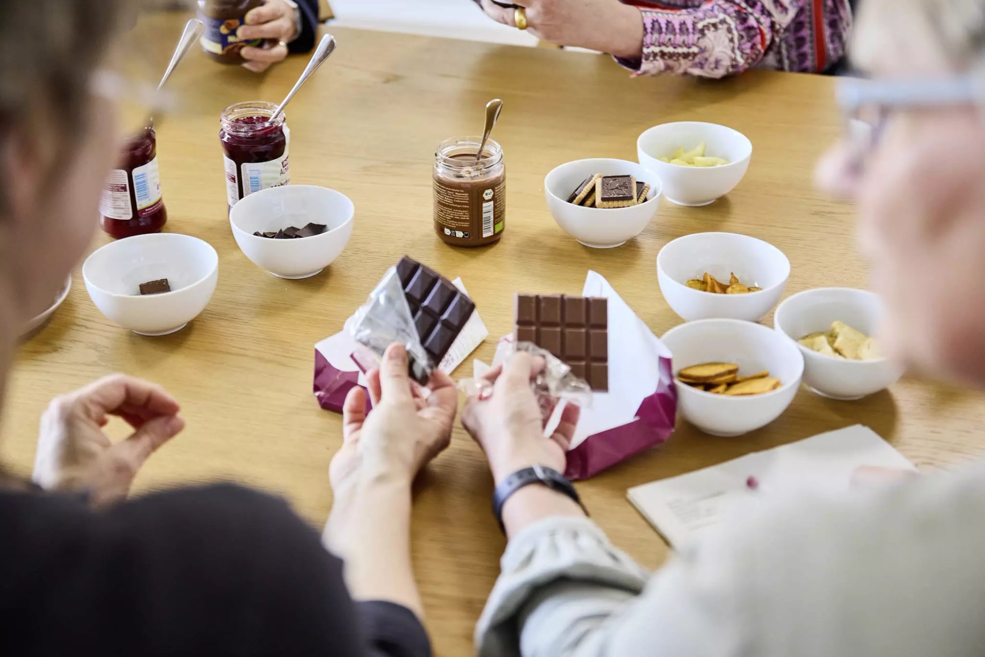 Mehrere Personen sitzen an einem Tisch mit Schalen voller Snacks und halten jeweils eine Tafel Schokolade in der Hand
