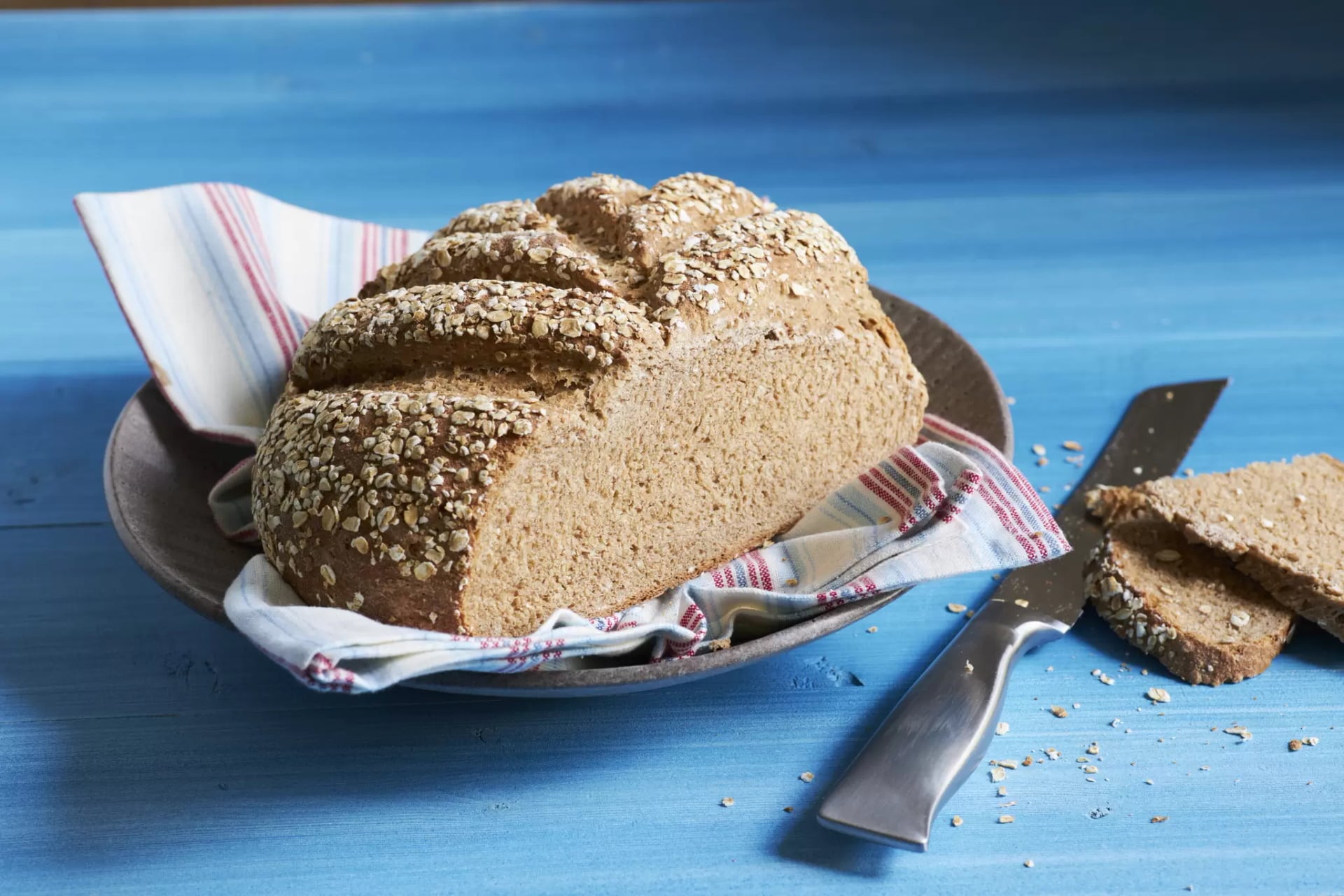Gebackenes Vollkornbrot mit Haferflocken auf einem Teller mit Stoffserviette und Brotmesser auf blauem Untergrund.