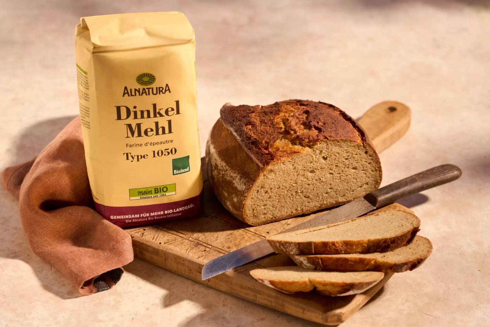 Eine Packung Dinkelmehl Typ 1050 neben einem angeschnittenen Kartoffelbrot mit mehreren Scheiben auf einem Holzbrett