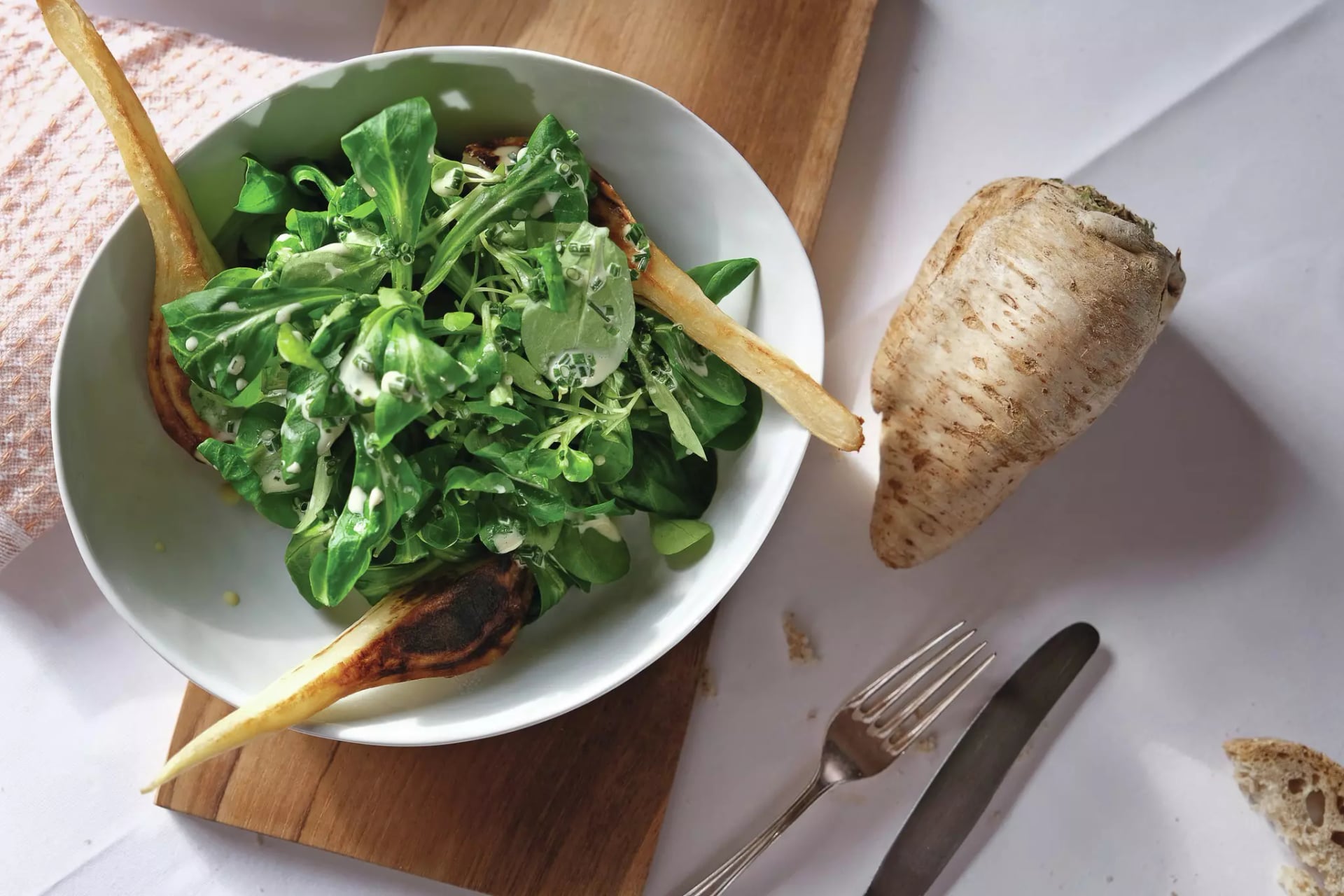Schüssel mit grünem Salat und aufgeschnittener gegrillter Pastinake auf einem Holzbrett daneben liegt eine Pastinake, eine Gabel und ein Messer. Ein Stück Brot ist am Rand auf weißer Tischdecke.
