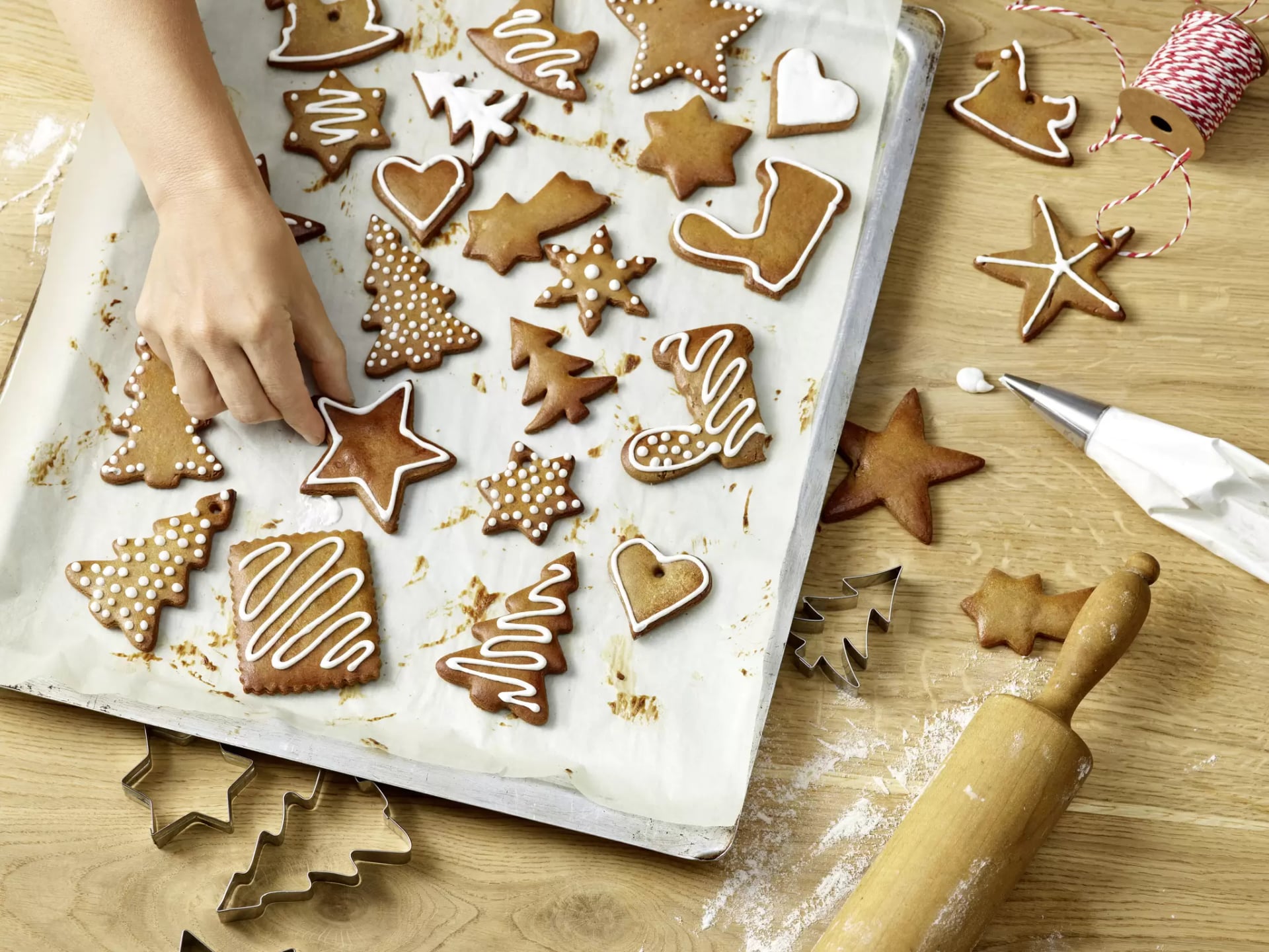 Lebkuchen in verschiedenen Formen liegen auf einem mit Backpapier ausgelegten Blech. Die Lebkuchen sind mit weißem Zuckerguss verziert, zum Beispiel als Sterne, Tannenbäume, Herzen und Stiefel. Eine Hand greift nach einem der Lebkuchen. Auf dem Holztisch daneben liegen Ausstechformen, ein Nudelholz, etwas Mehl und eine Spritztüte mit weißem Zuckerguss. Im Hintergrund ist eine Rolle mit rot-weißem Bäckergarn zu sehen.