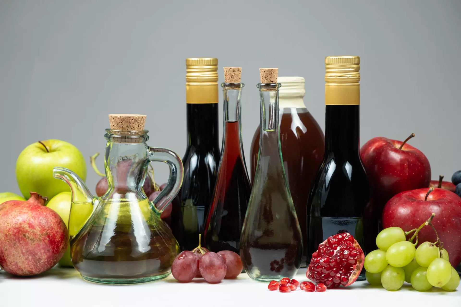 Essige in verschiedenen Sorten und Flaschen auf einer weißen Platte umgeben von Obsorten wie Äpfeln, Trauben und Granatapfel.