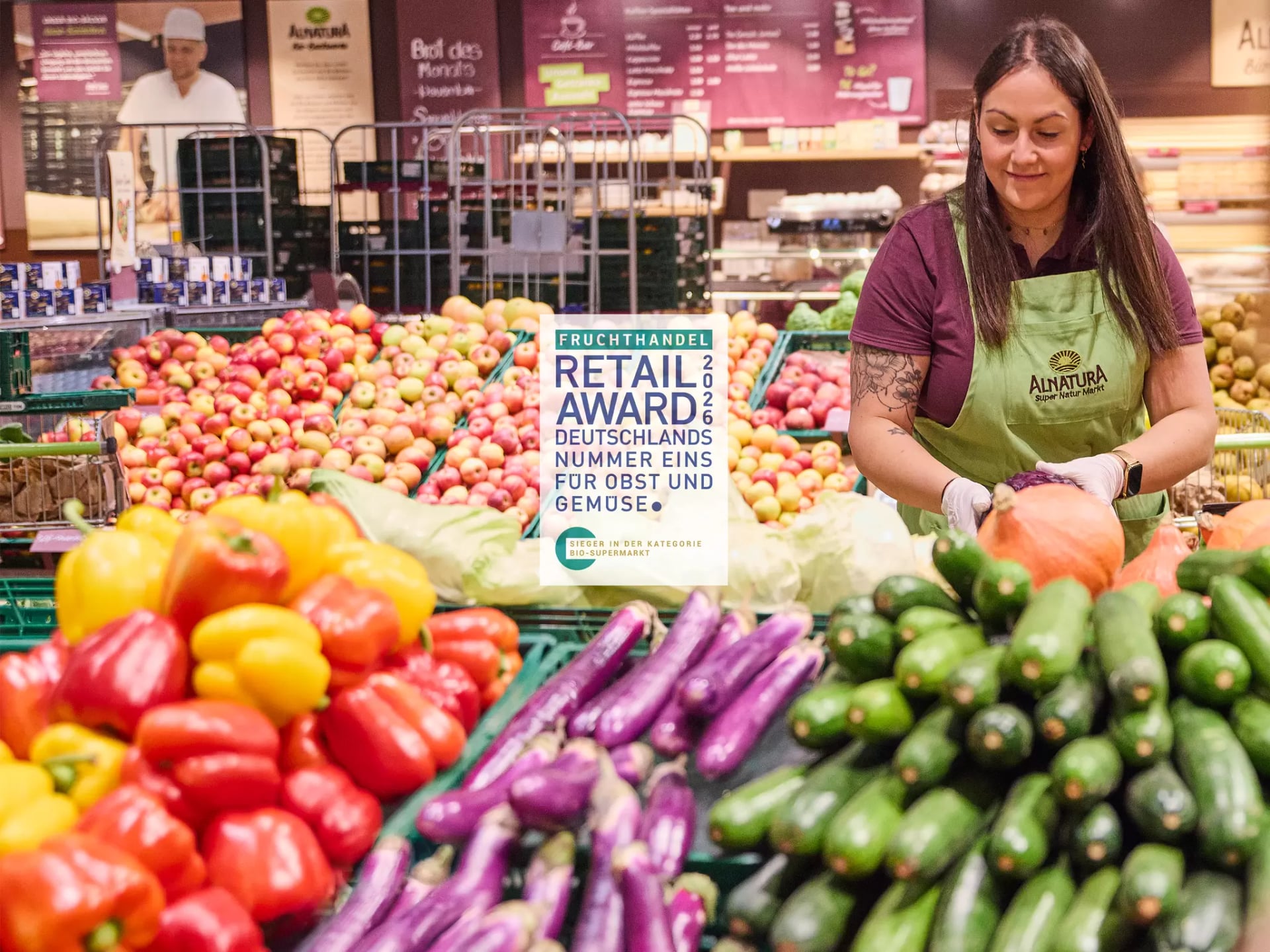 Alnatura Mitarbeiterin verräumt Ware am Obst- und Gemüseaufbau in einem Alnatura Markt. In der Mitte das Logo des Retail Awards mit dem Text: "Fruchthandel Retail Award 2026 - Deutschlands Nummer Eins für Obst und Gemüse. Sieger in der Kategorie Bio-Supermarkt.