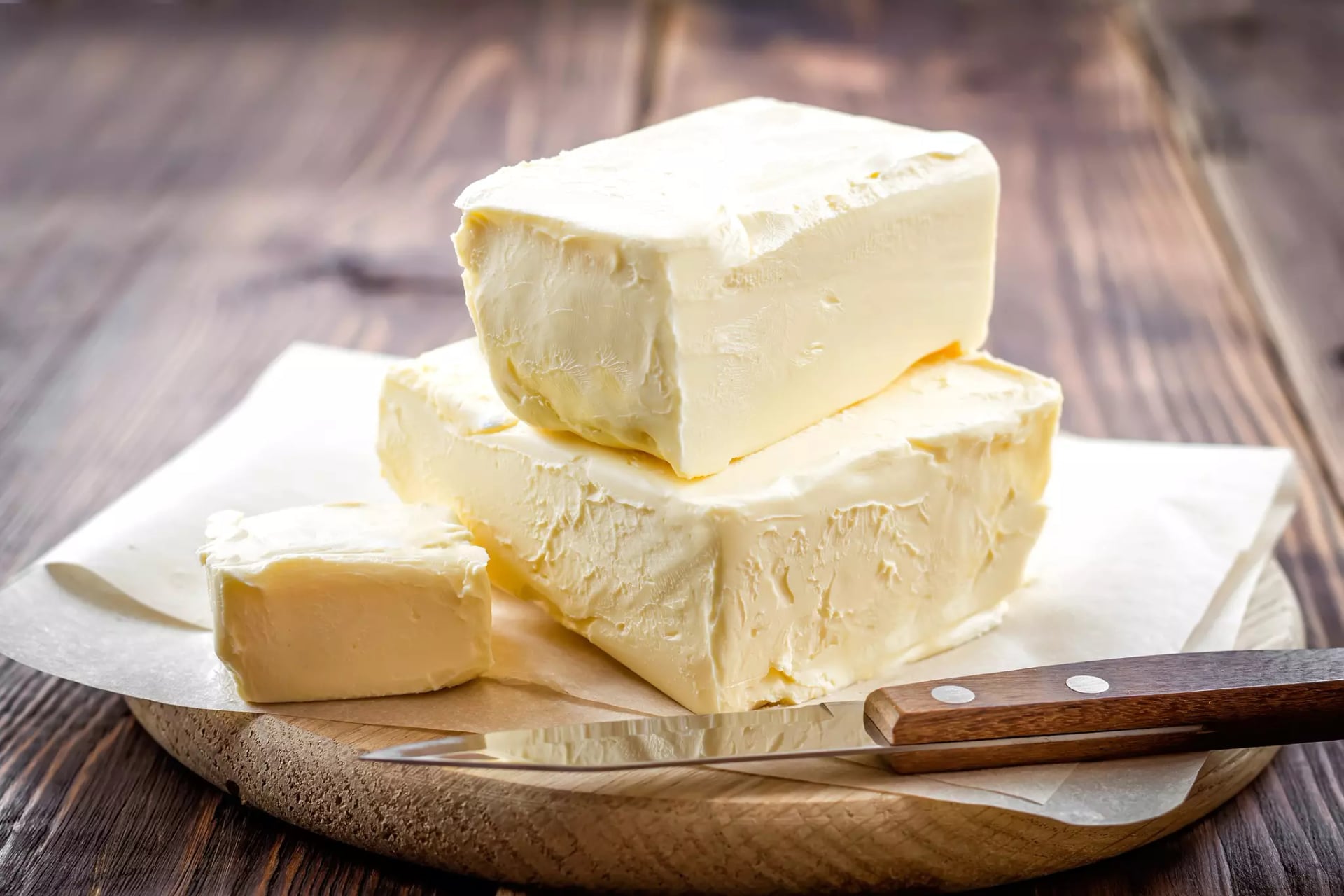 Zwei weich aussehende Butterblöcke aufeinandergestapelt auf Butterpapier, das auf einem Holzbrett liegt. Ein Block wurde mit einem Messer angeschnitten.