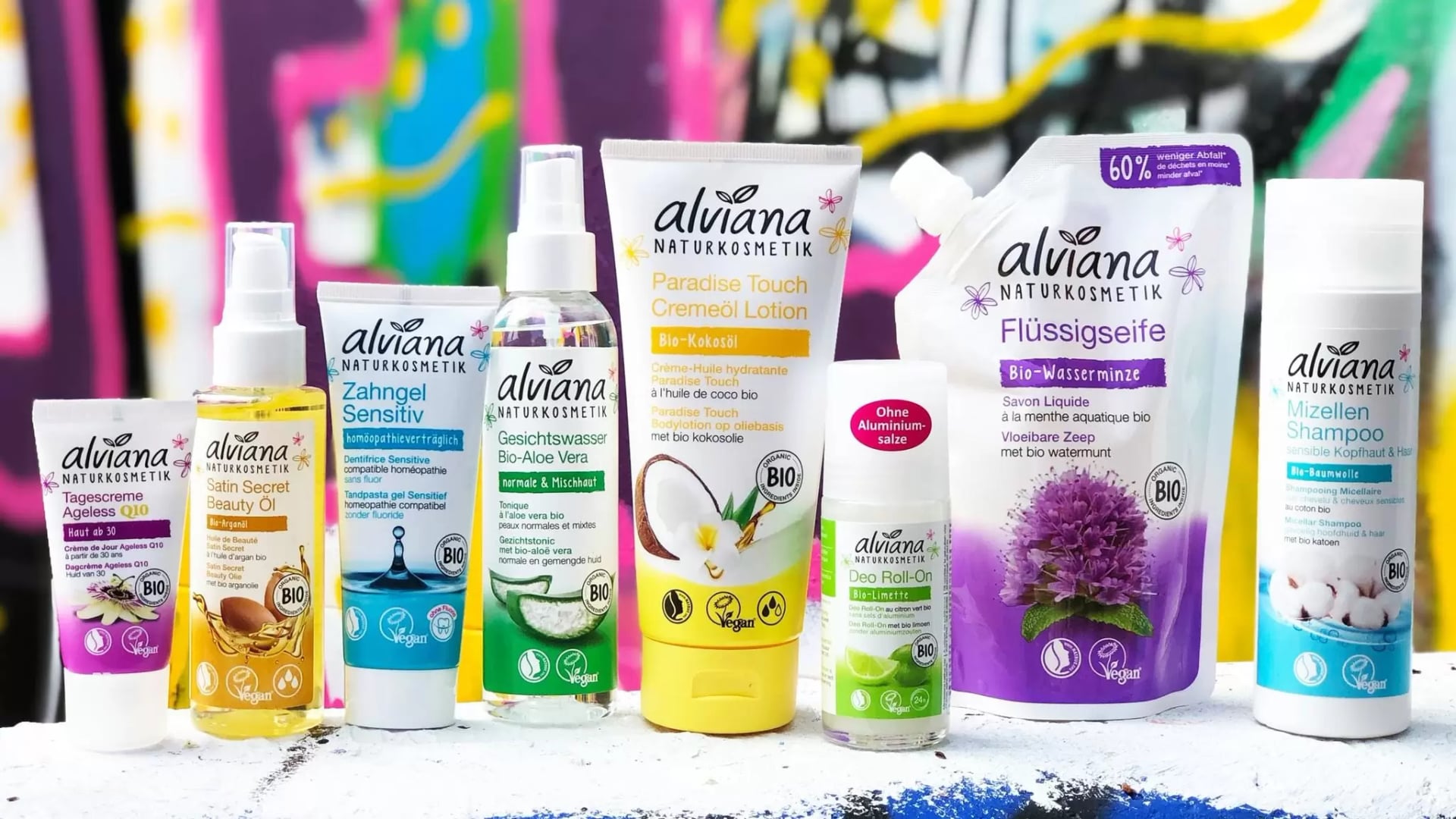 Acht verschiedene alviana Naturkosmetik-Produkte in weißen Verpackungen mit bunten Etiketten stehen vor einer bunten Graffiti-Wand