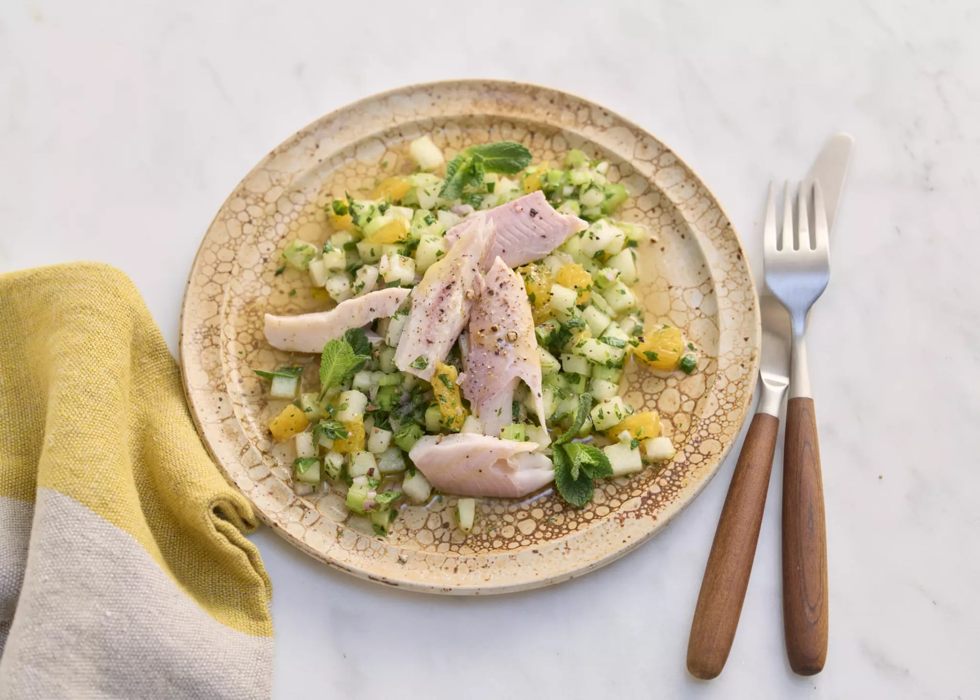 Teller mit Forellenfilets auf einem Melonensalat aus gewürfelter Melone und Gurke, garniert mit Kräutern, daneben Besteck mit Holzgriffen und ein gelb-beiger Stoffserviette.
