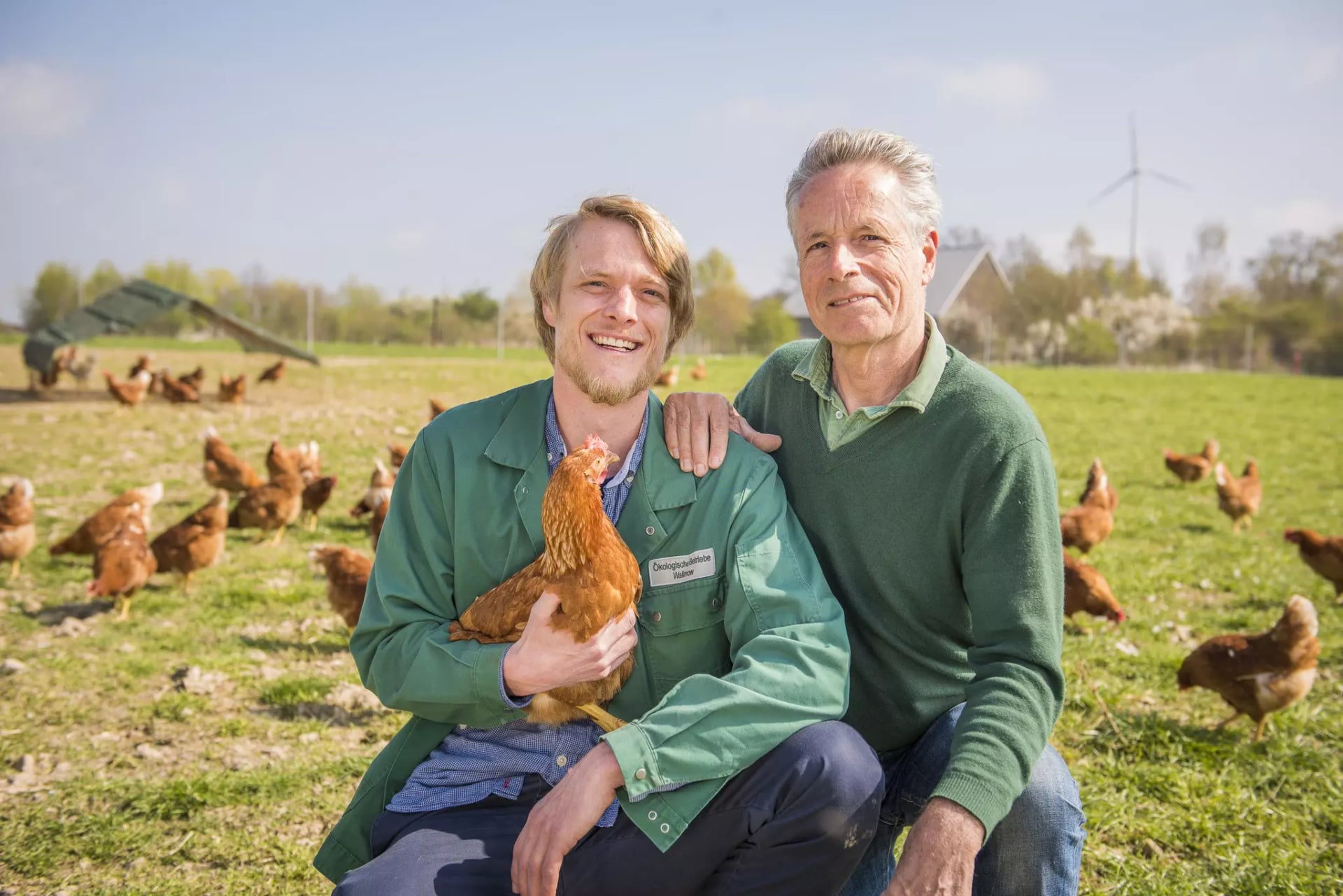 Zwei Männer sitzen auf einer Wiese mit freilaufenden Hühnern, der jüngere hält ein Huhn im Arm, der ältere legt die Hand auf dessen Schulter.