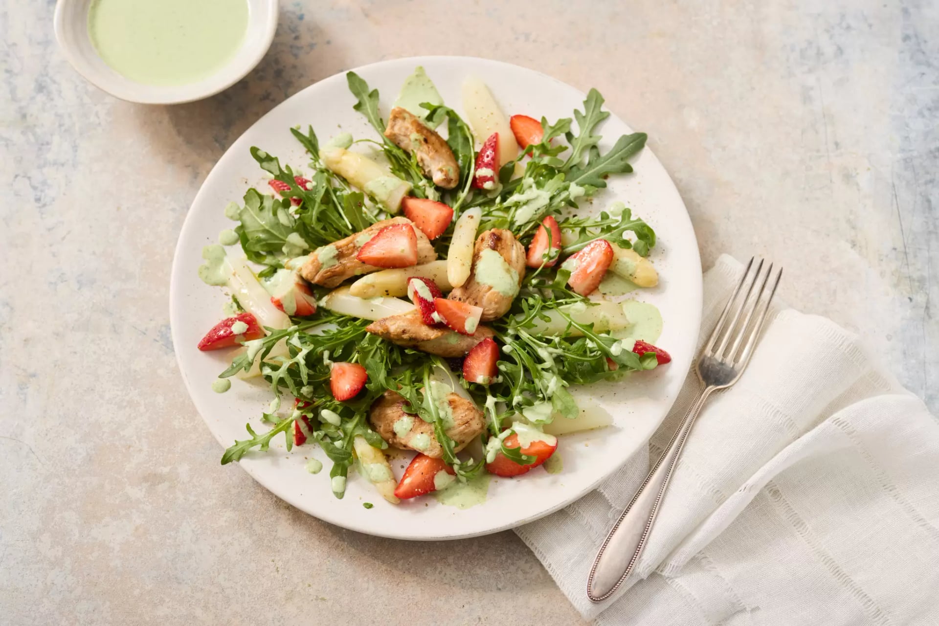 Teller mit Erdbeer-Spargel-Salat aus grünem Salat, weißen Spargelstücken, Erdbeeren und gebratenen Hähnchenstreifen, daneben eine Gabel auf einem weißen Stoffserviette und eine kleine Schale mit Dressing.