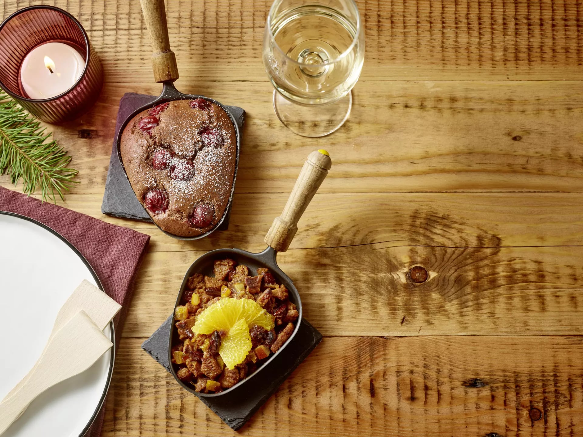 Zwei kleine Pfannen mit gebackenem Dessert, eine mit Puderzucker und Beeren, die andere mit Orangenscheiben, auf einem Holzbrett neben einem Glas Weißwein, einer Kerze und einem Teller mit Serviette.