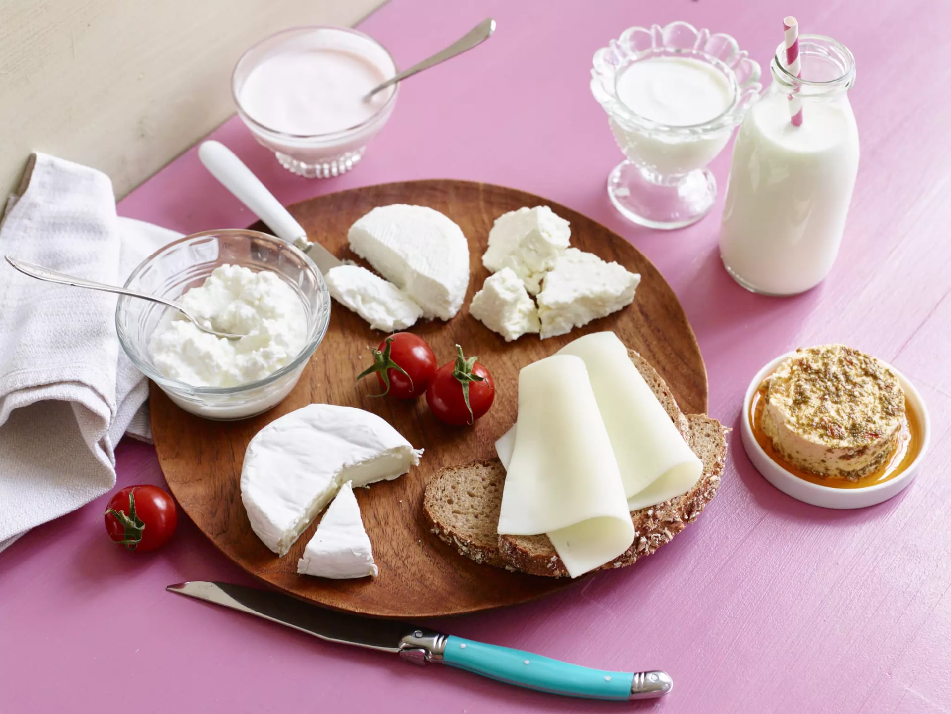 Holzbrett mit verschiedenen Käsesorten, Tomaten, Vollkornbrot, Joghurt in Gläsern und einer Flasche Milch auf rosa Untergrund