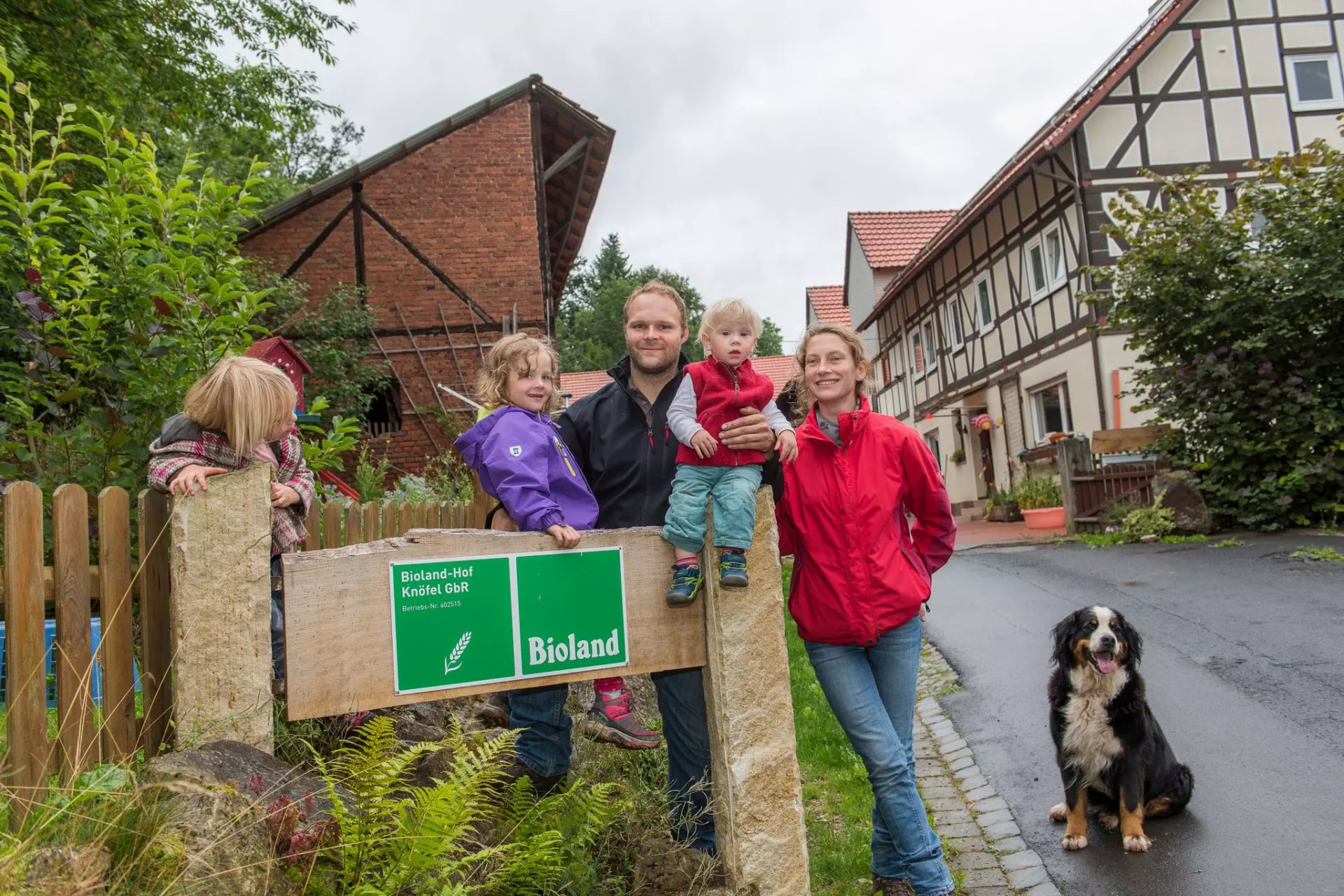 Bild der Familie Knoefel am Eingang zu ihrem Hof. Rechts der Familienhund. Zwei Erwachsene und drei Kinder. Im Hintergrund das Wohnhaus (Fachwerk).