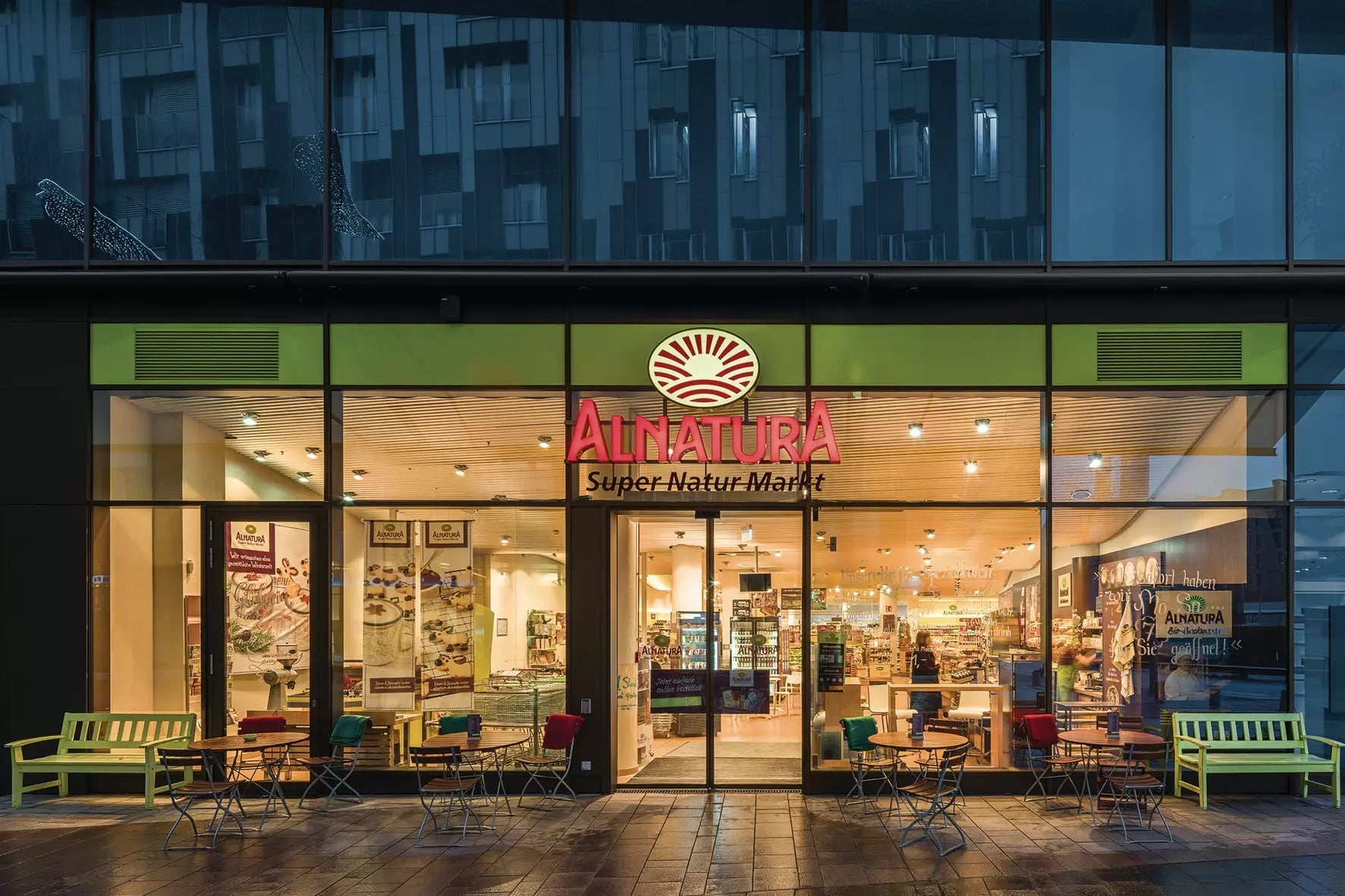Außenansicht des Alnatura Super Natur Markts Hamburg-Hafencity mit beleuchtetem Eingangsbereich und Sitzgelegenheiten davor.