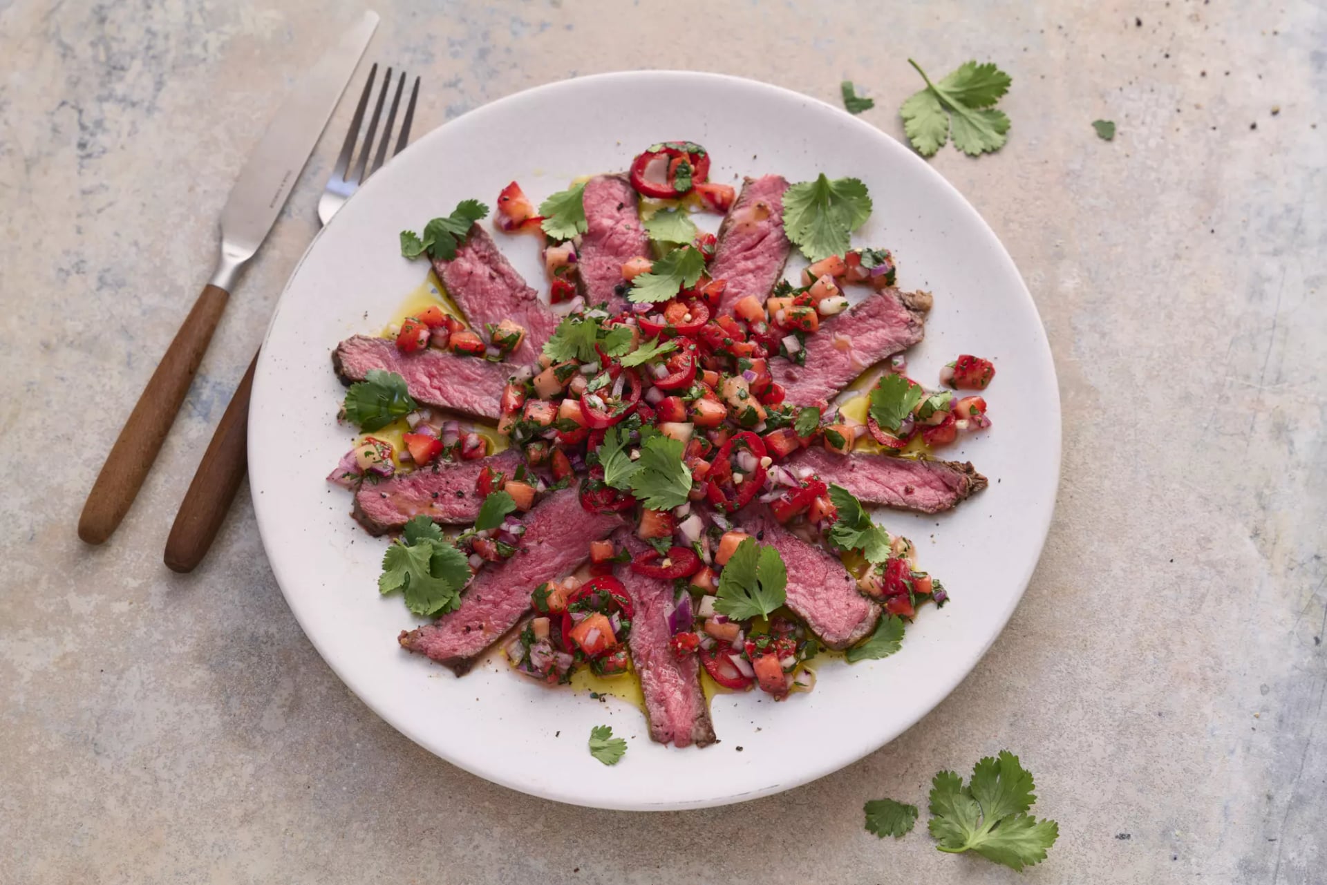 Auf einem weißen Teller sind dünne Scheiben von rosa gebratenem Rostbeef mit einer Erdbeer-Salsa und Korianderblättern angerichtet, daneben eine Gabel und ein Messer mit braunen Griffen auf einer hellen Oberfläche.