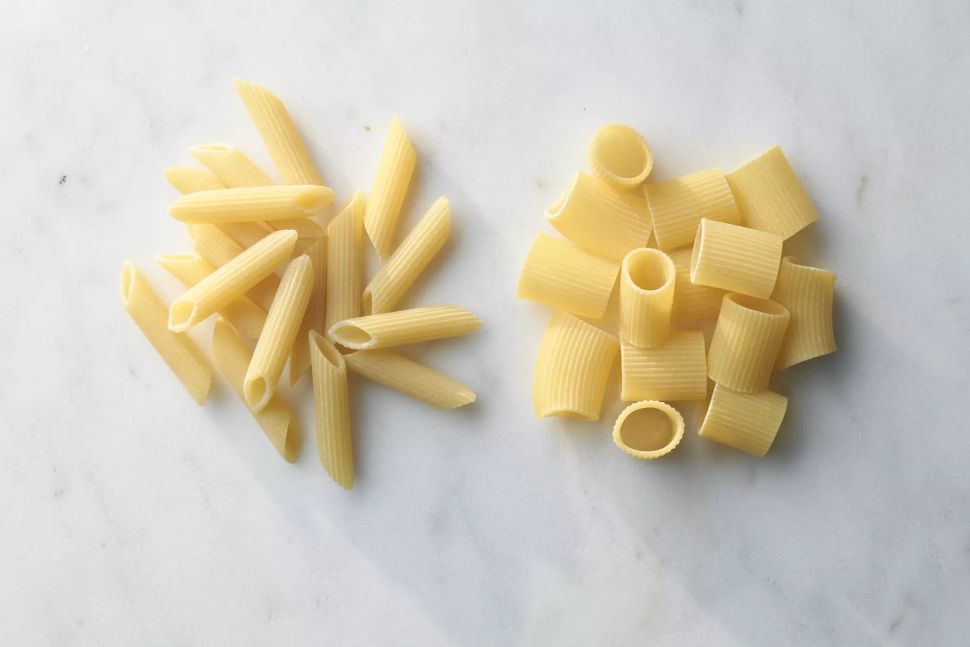 Zwei Sorten ungekochte Pasta nebeneinander auf hellem Untergrund, links Penne, rechts Rigatoni