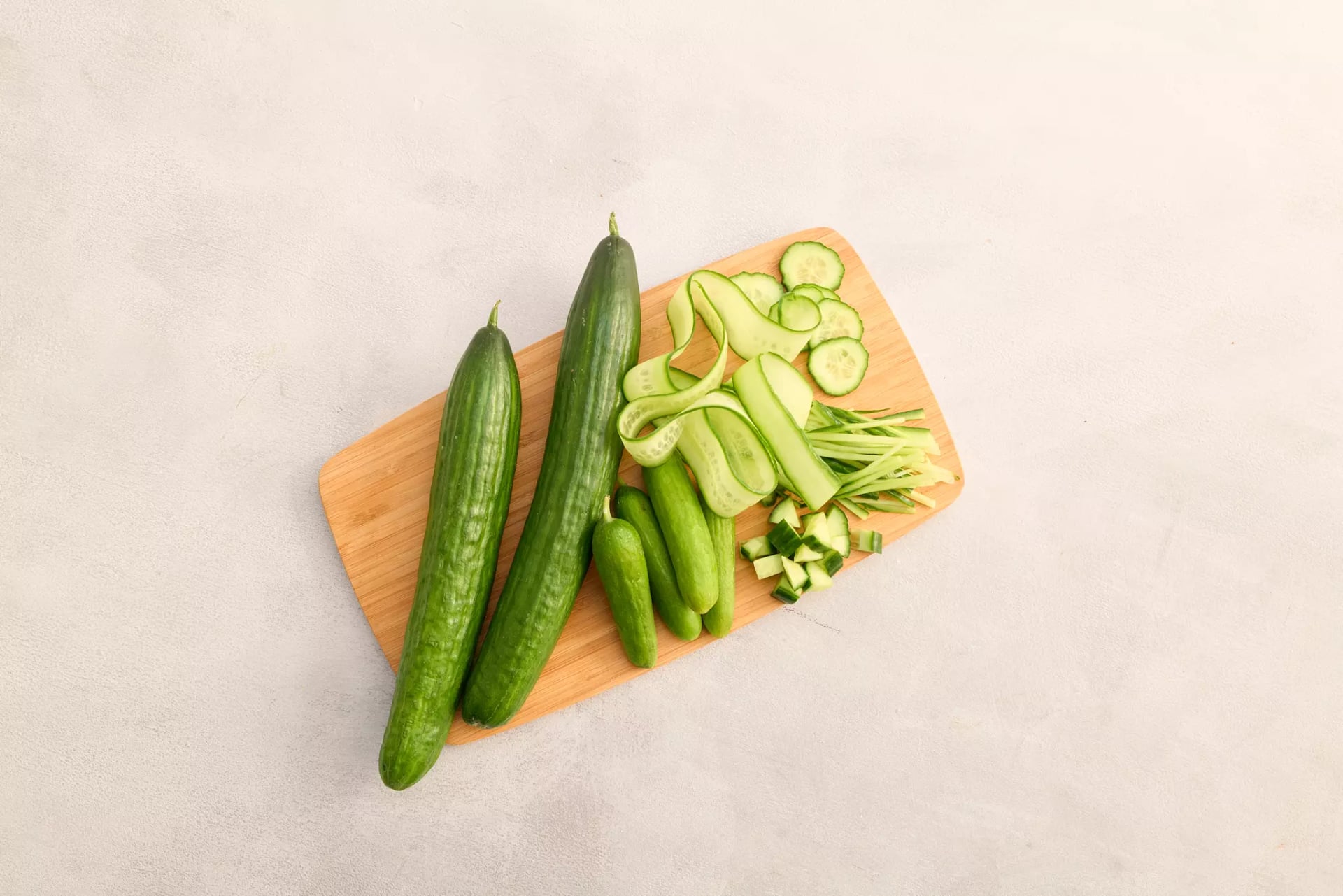 Auf einem hellen Grund liegt ein Holzbrett mit Salatgurken und Snackgurken. Eine Gurke wurde verschieden aufgeschnitten, in Scheiben, Streifen und gewürfelt.