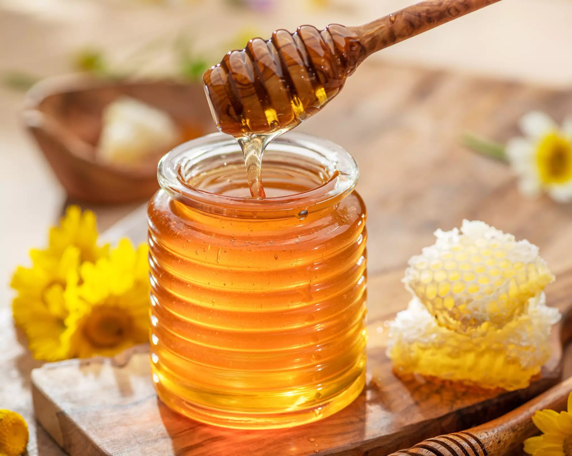 Honig in einem geriffelten Glas wird mit einem Holz-Honiglöffel angehoben. Daneben Honigwaben und gelbe Blüten.