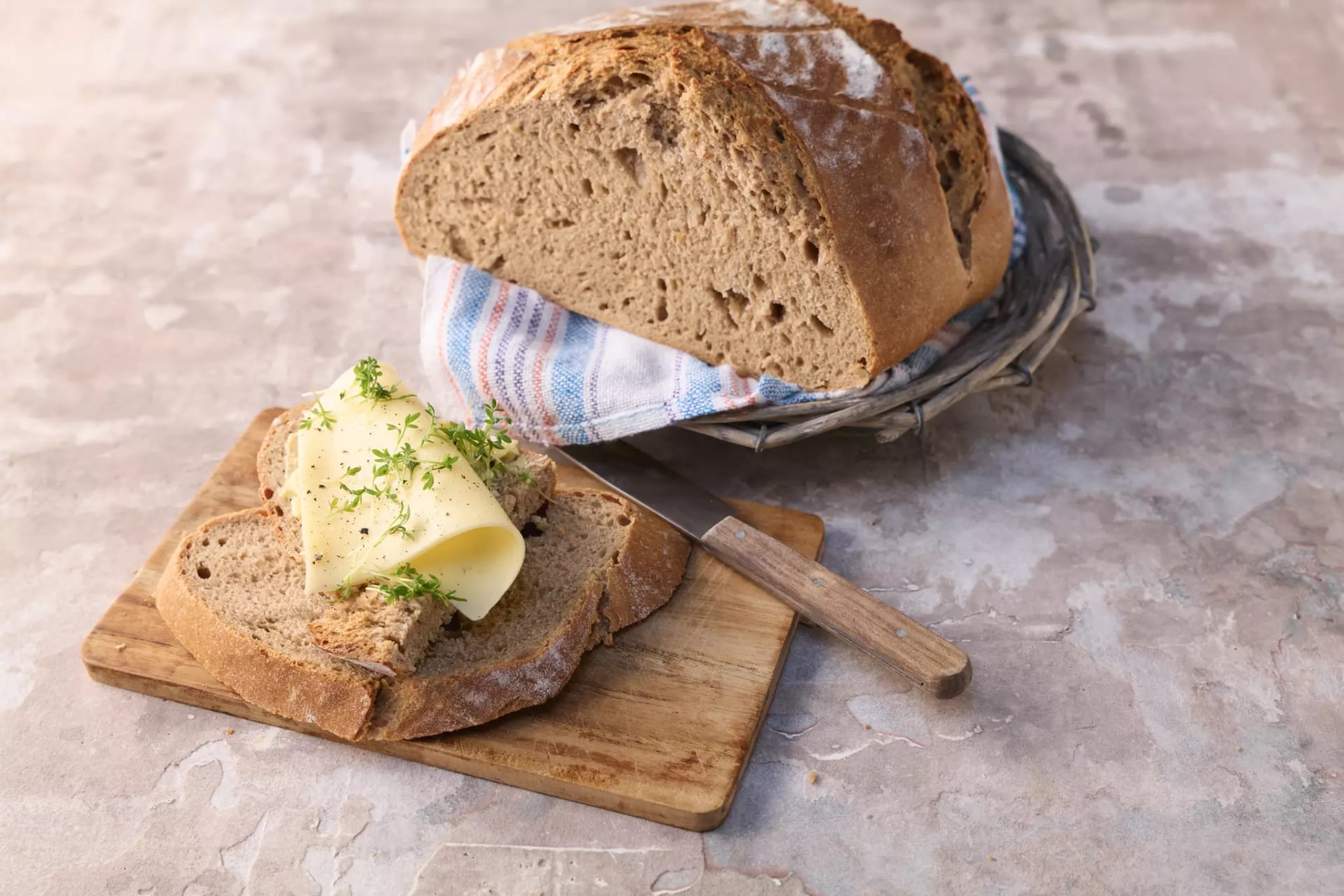 Knuspriges Dinkel-Sauerteigbrot in einem Brotkorb auf einem hellen, strukturierten Untergrund, davor ein Holzbrett mit einer Scheibe Brot, belegt mit Käse und frischen Kräutern, daneben ein Messer mit Holzgriff.