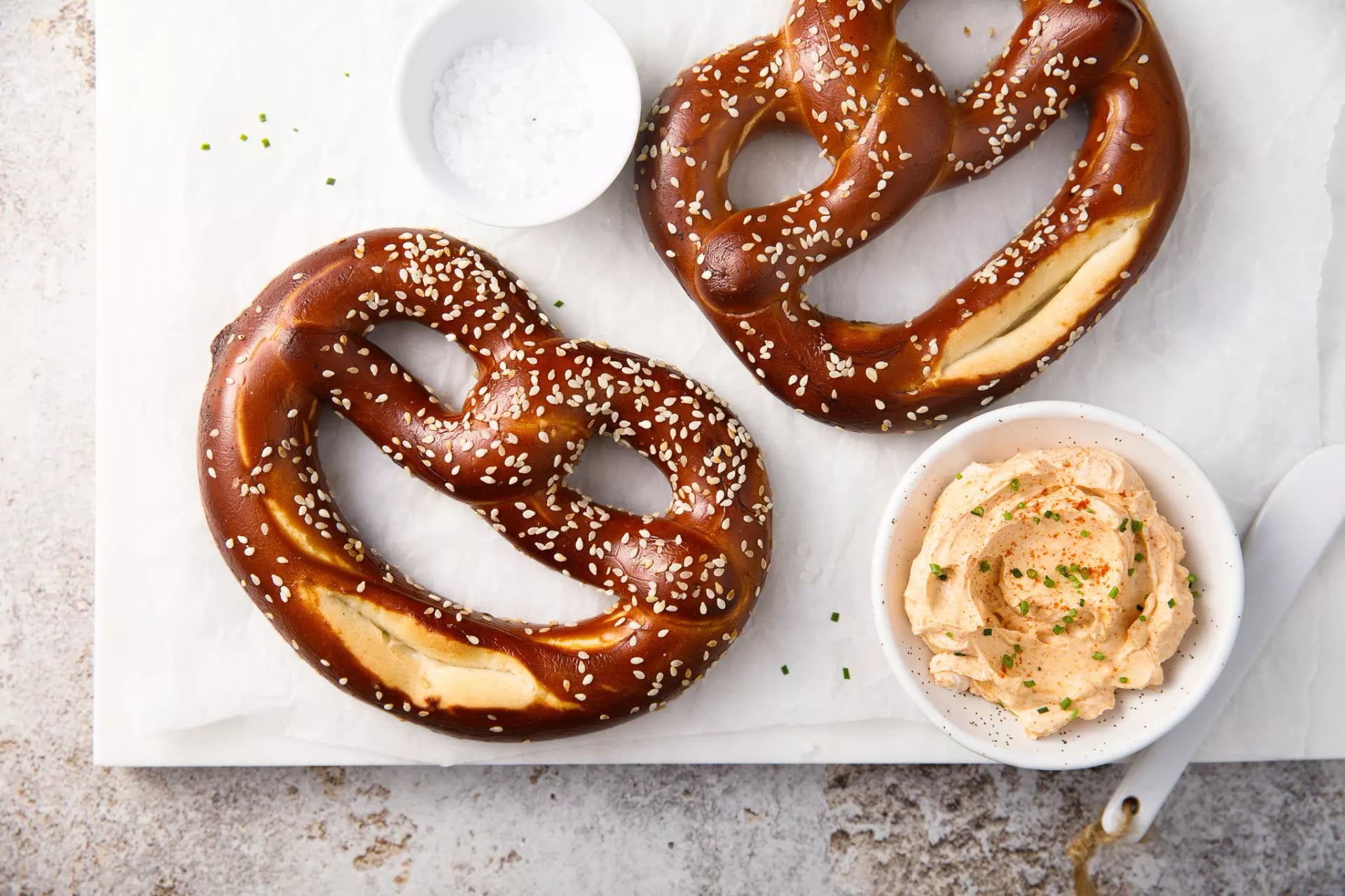 Zwei Laugenbrezeln mit Sesam liegen auf einer weißen Platte, daneben eine Schale mit Salz und eine Schale mit einem Dip, der Spundekäs ähnelt.