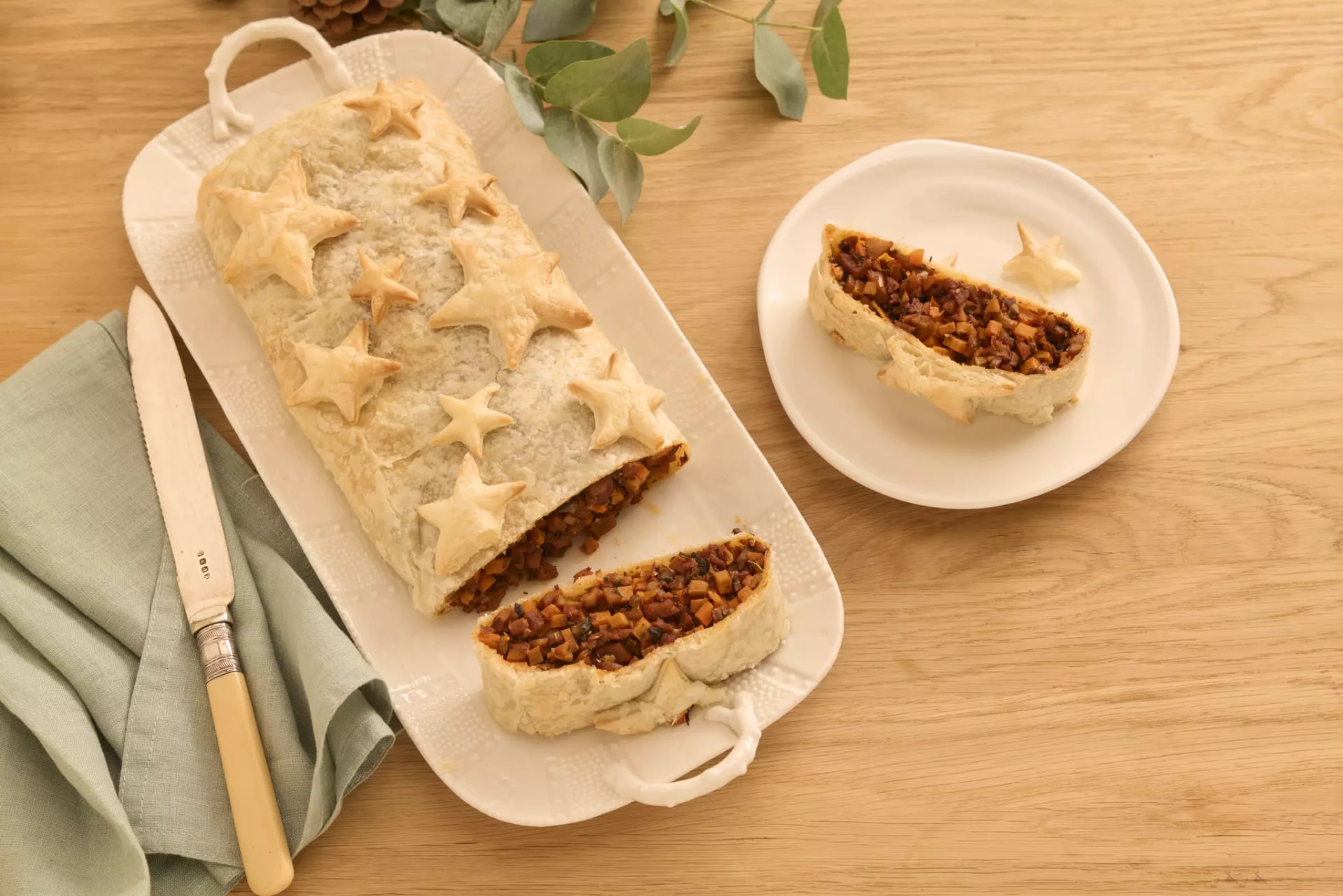 Auf einer weißen rechteckigen Porzellanplatte ist ein goldbraun gebackener Strudel, dieser ist gefüllt mit einer Mischung aus Seitan, Süßkartoffelwürfeln und Pilzen. Eine Scheibe des Strudels ist abgeschnitten und auf einem weißen Teller angerichtet, eine andere Scheibe liegt noch auf der weißen Platte. Die Füllung ist deutlich sichtbar, und die Oberfläche ist knusprig gebacken. Neben der Platte ist noch ein Messer und eine türkisfarbene Stoffserviette zu sehen.