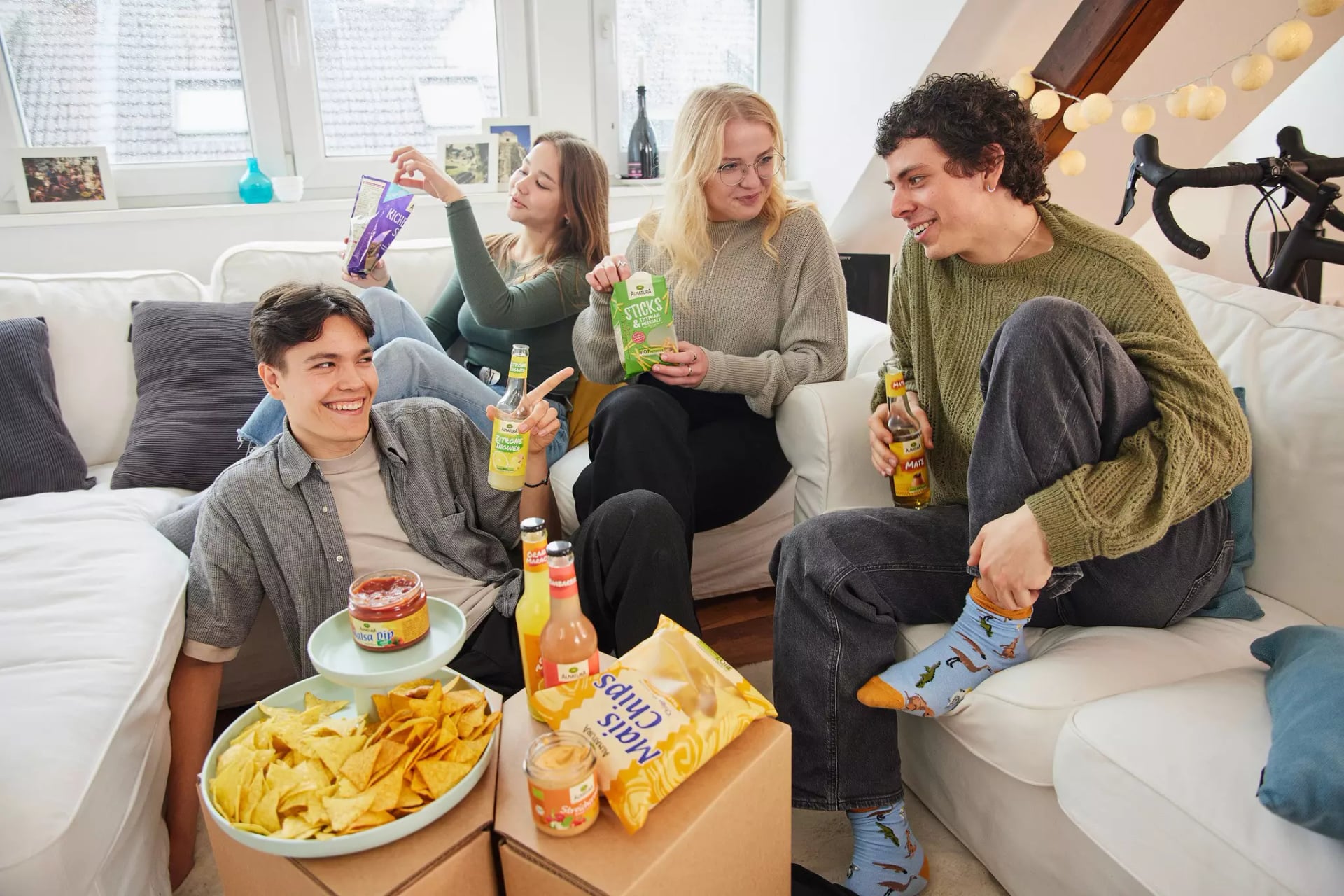 Vier junge Menschen sitzen auf Sofas in einem Wohnzimmer und essen Chips und trinken Getränke
