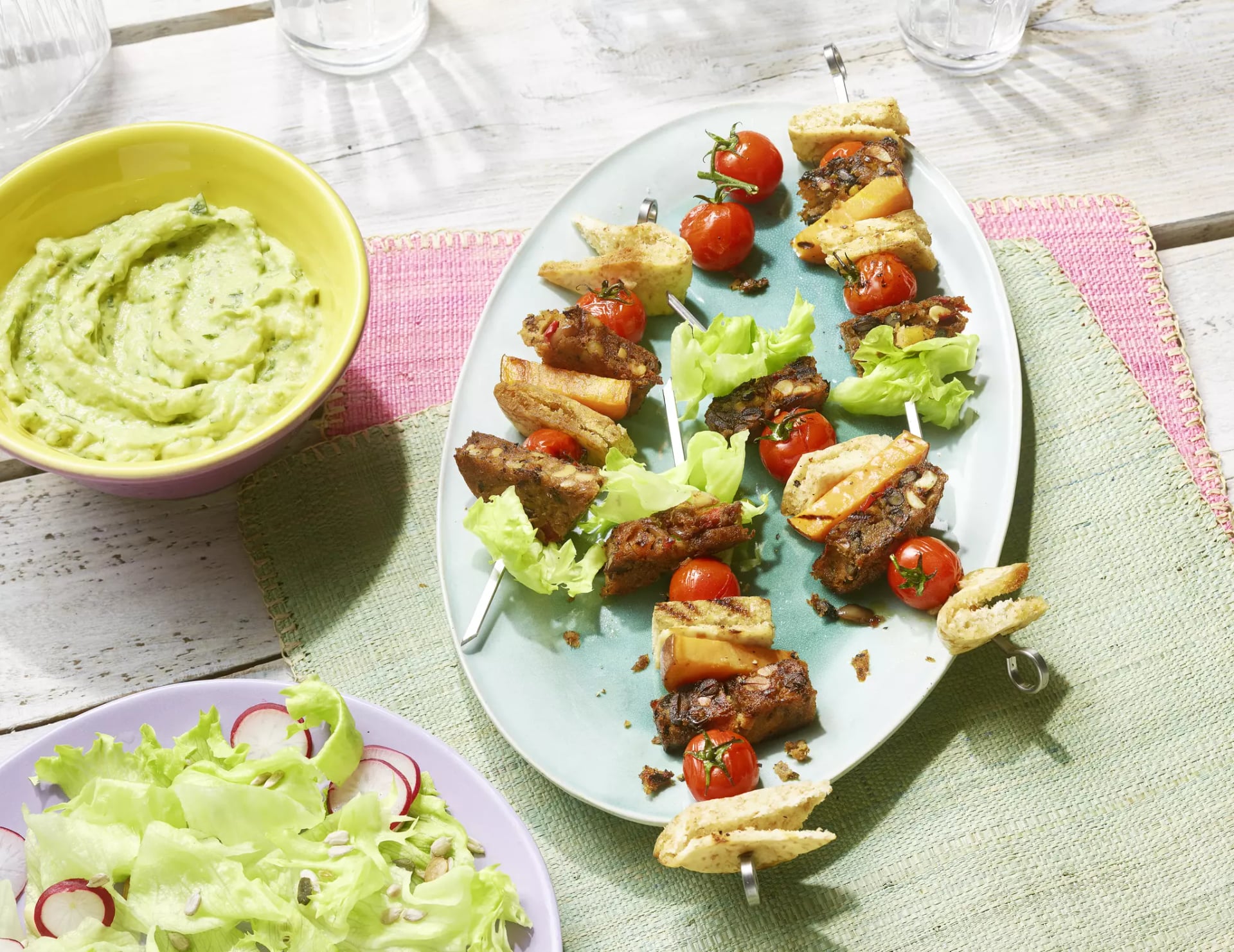Spieße mit Fleisch, Tomaten, Salat und Brot auf einem Teller neben einer Schüssel mit grüner Creme und einem Salat mit Radieschen.
