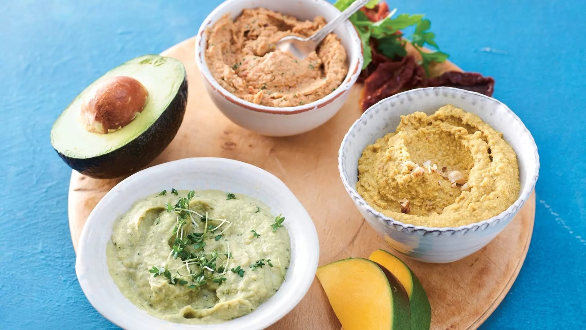 Drei Schalen mit verschiedenen Hummus-Sorten auf einem runden Holzbrett daneben eine halbierte Avocado und Mangostücke