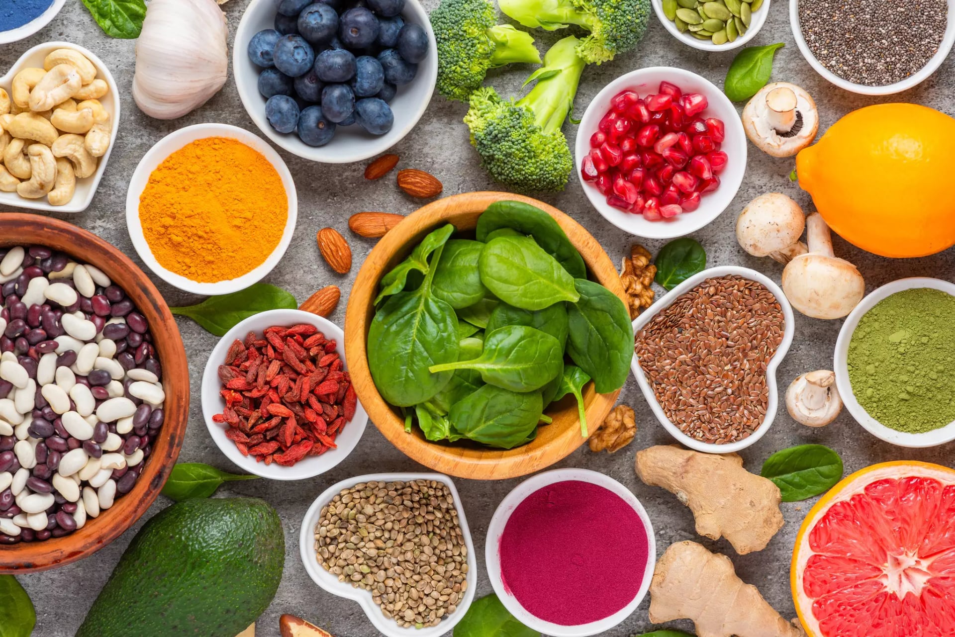 Eine bunte Zusammenstellung verschiedener Superfoods wie Spinat, Gojibeeren und Leinsamen angerichtet - teilweise in Schalen - auf einem betonartigen Untergrund