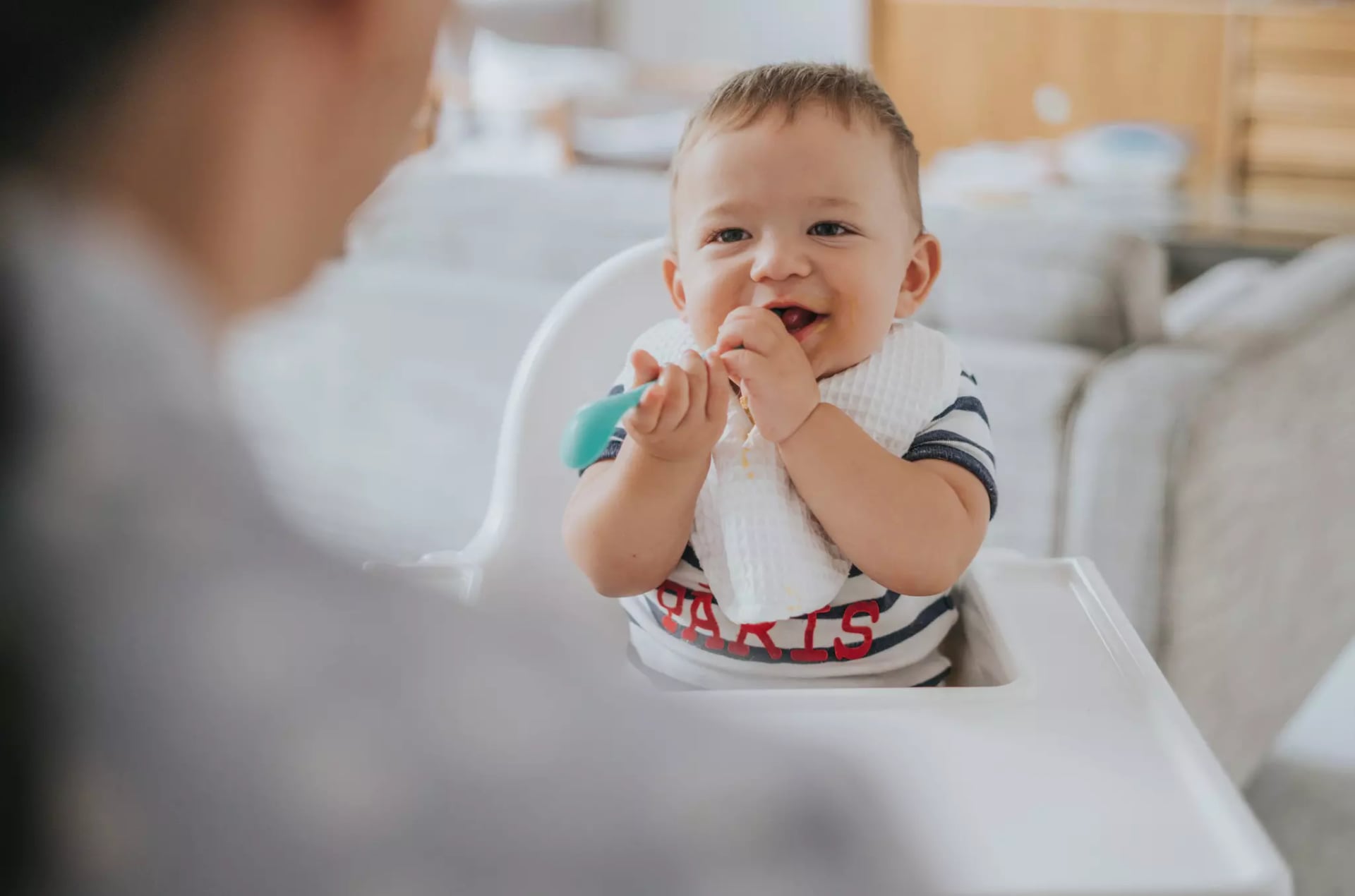 Ein Baby sitzt auf einem Hochstuhl und steckt sich lachend einen türkis farbenen Plastiklöffel falsch herum in den mit essen verschmierten Mund
