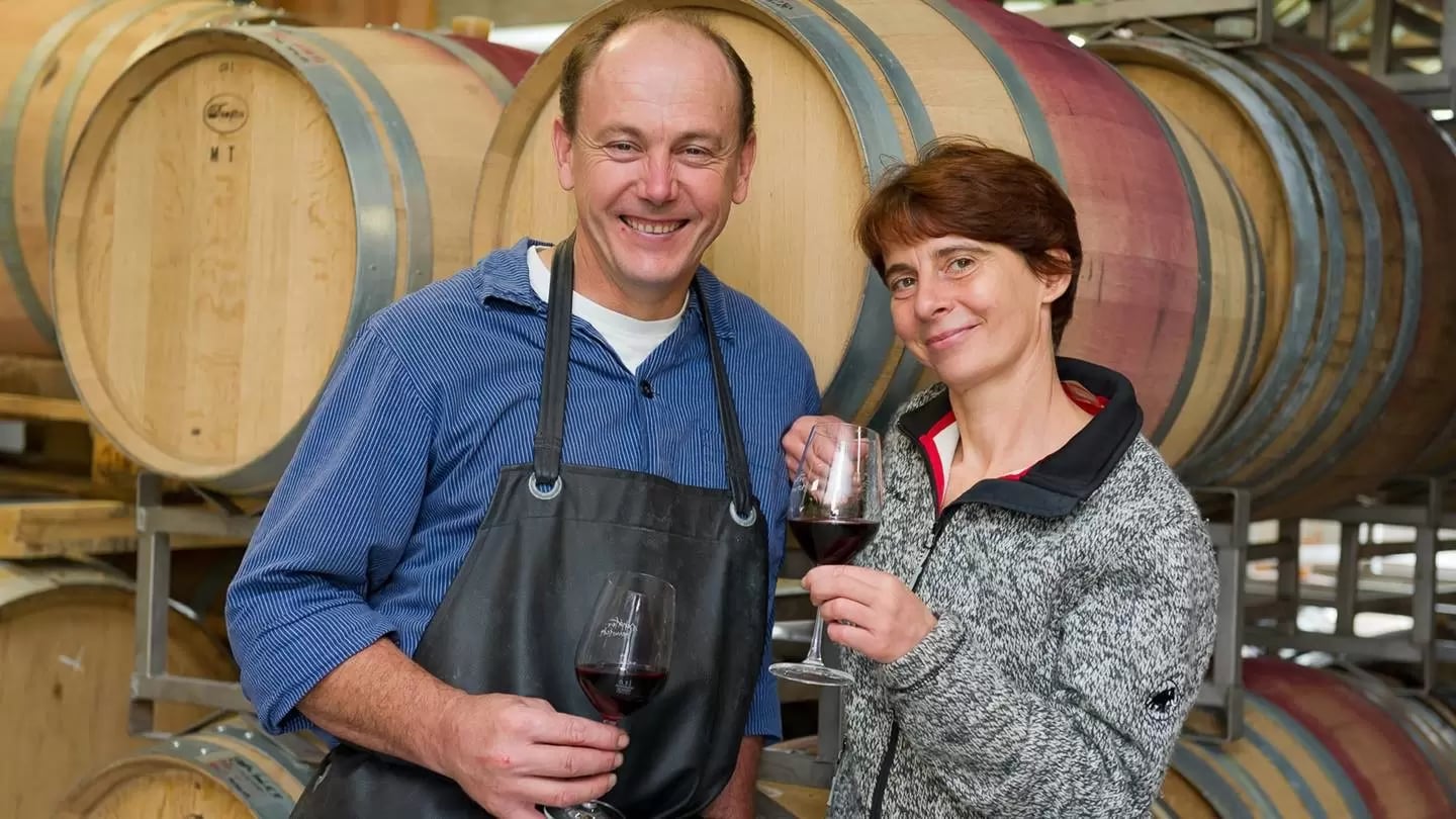 Ein Mann und eine Frau stehen vor mehreren gelagerten Holzfässern und halten jeweils ein Glas Rotwein in der Hand.